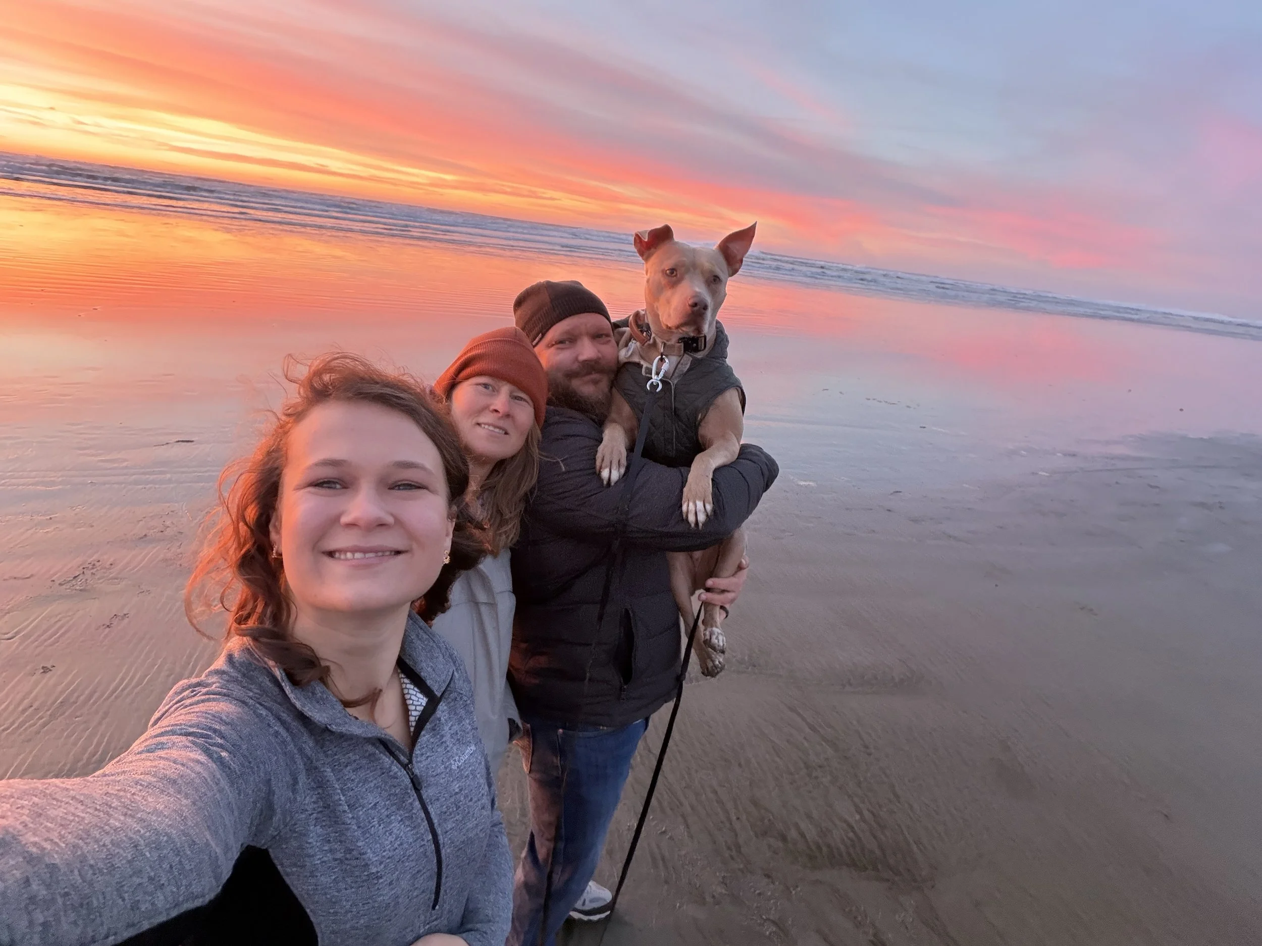 A group of four people and a dog taking a selfie on a beach at sunset with colorful sky and calm water.