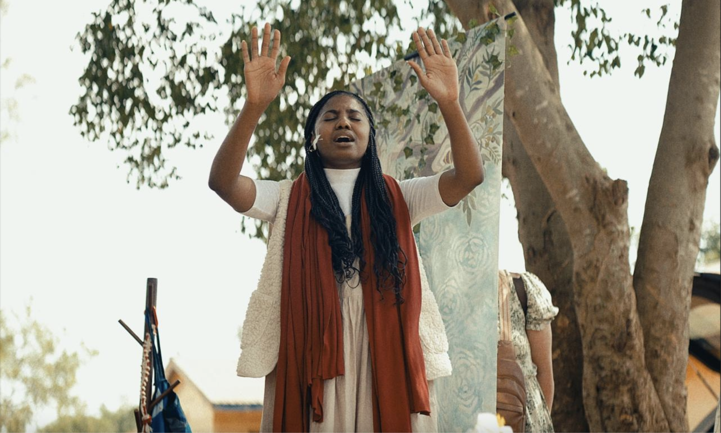 A woman with braided hair and wearing a white top, red scarf, and cream-colored vest is standing outdoors with her eyes closed and hands raised, appearing to be praying or singing introspectively. There are trees and a colorful mural or fabric behind her.
