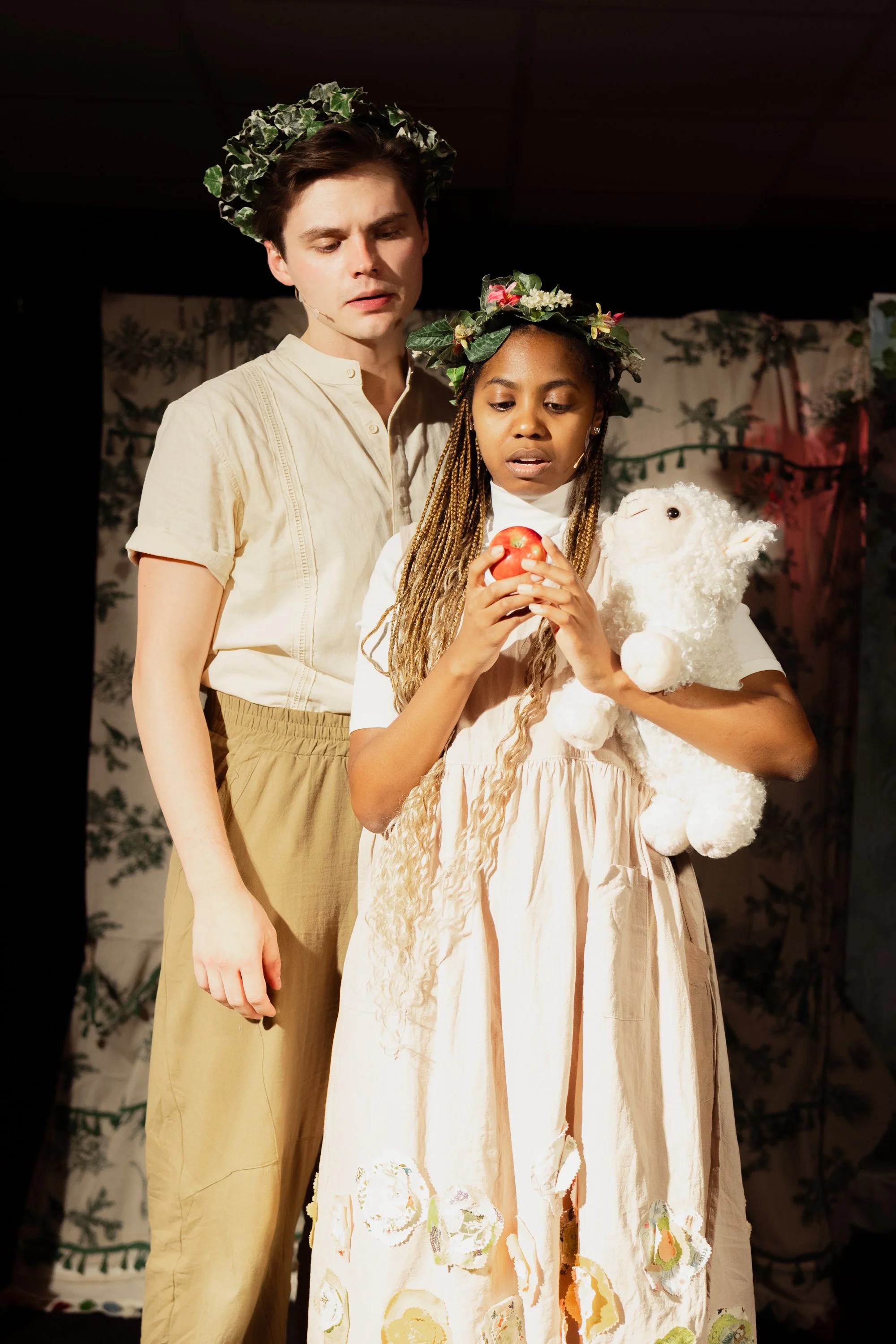 Two young performers on stage, wearing flower crowns, with one holding a stuffed animal and an apple, suggesting a theatrical or storytelling scene.