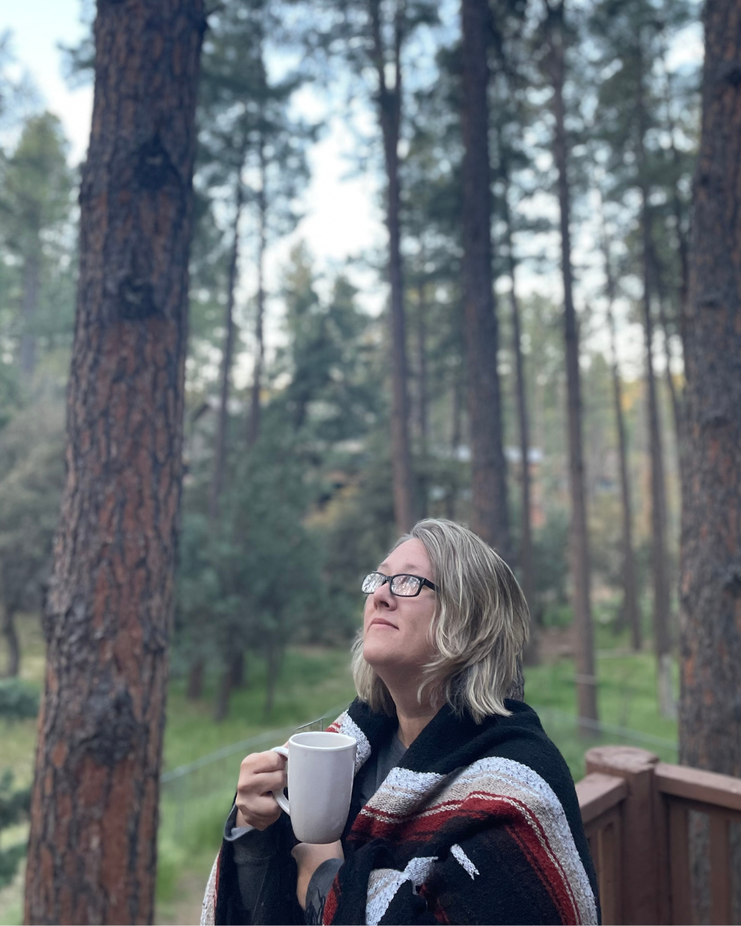 A woman with glasses and blonde hair, wrapped in a blanket, holding a white mug, standing on a wooden deck forested background with tall pine trees.