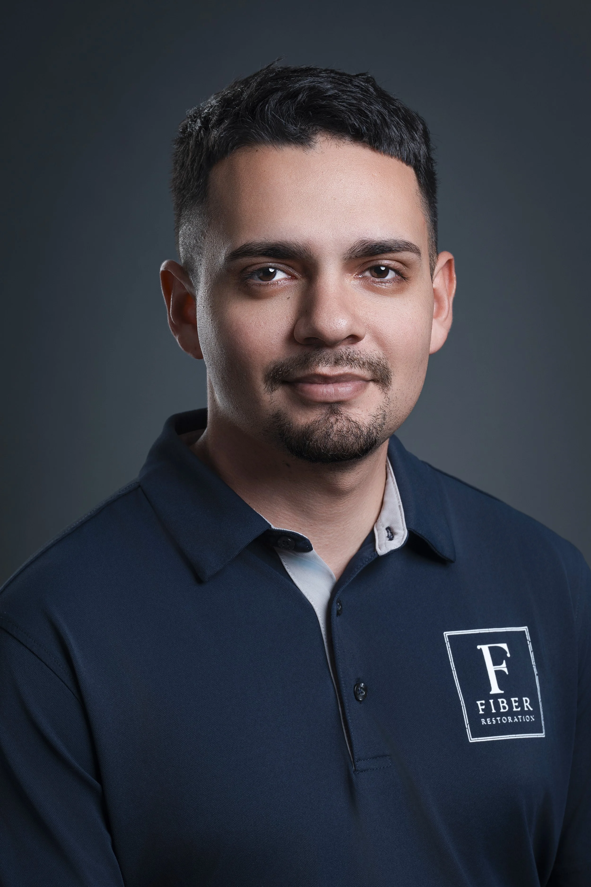 Portrait of a young man with short dark hair, stubble, wearing a navy blue polo shirt with the logo 'Fiber Restoration' on it, posing against a dark gray background.