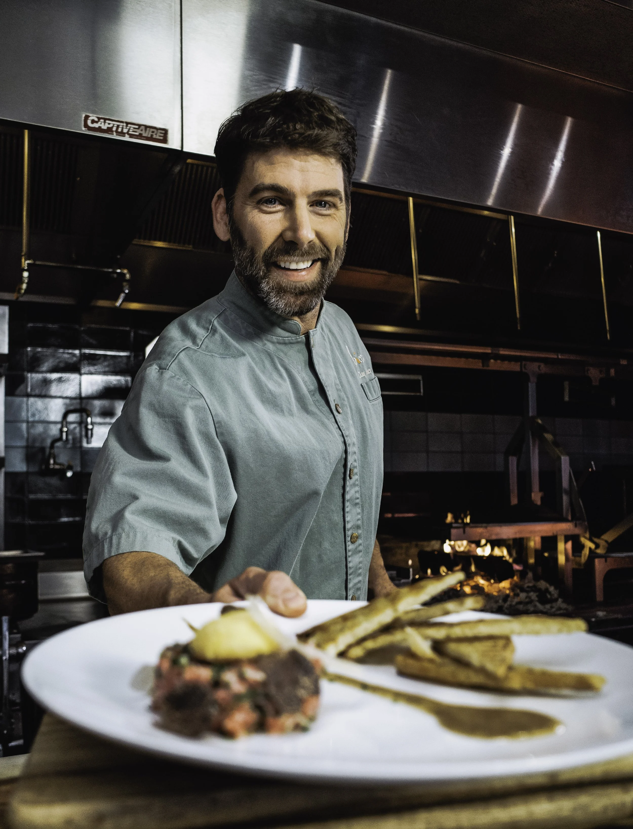 A smiling chef presenting a plate with beef, potato, and fried chips in a commercial kitchen.