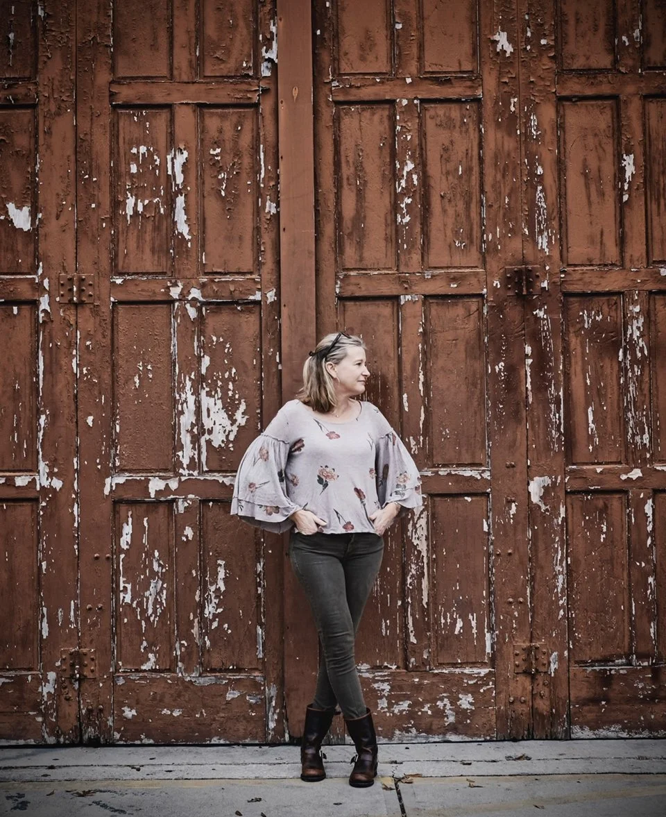 A woman standing in front of a large, rustic, brown wooden door with peeling paint, wearing a gray floral blouse, black jeans, and black boots.