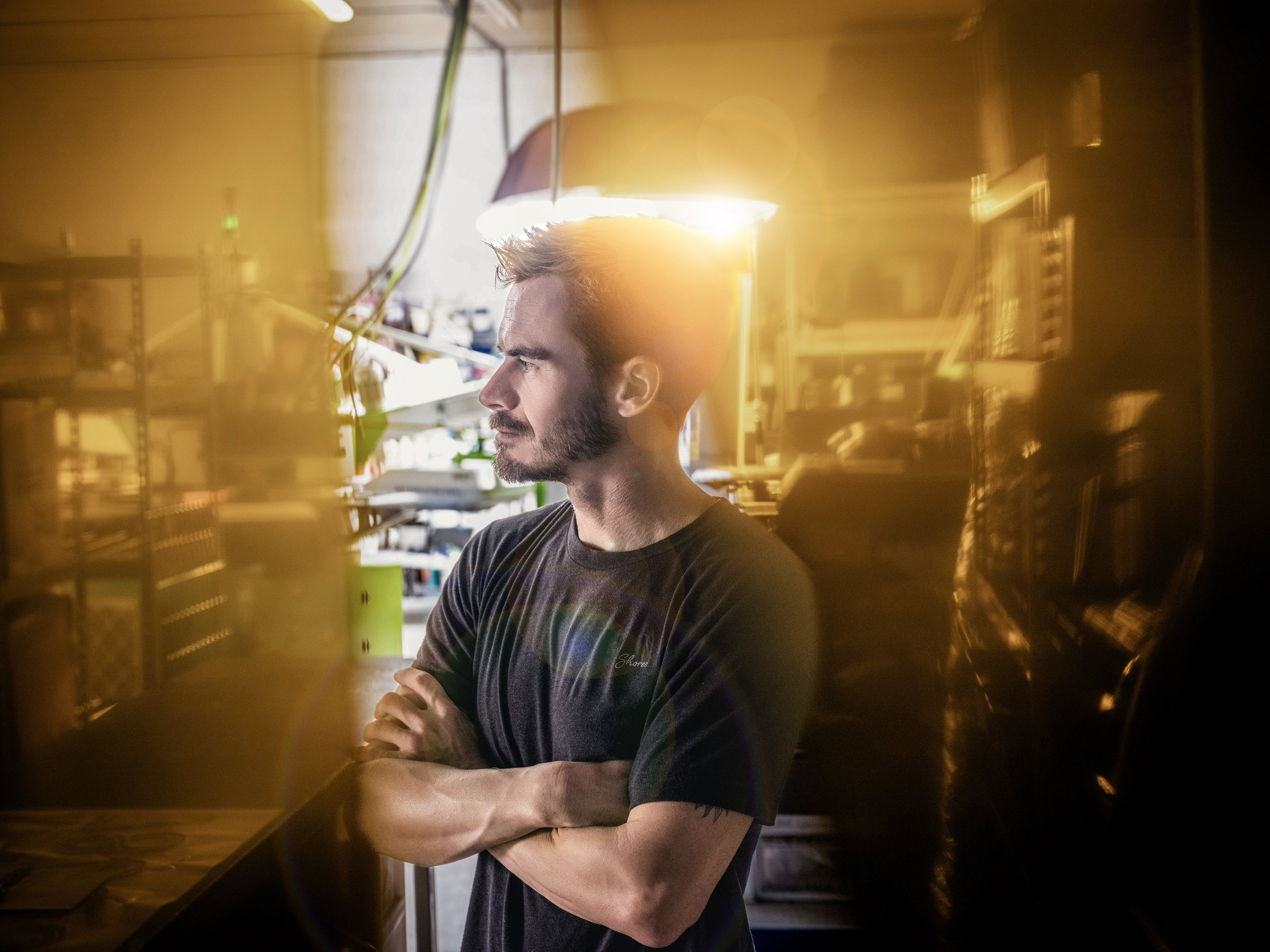 Bearded man in a black T-shirt standing with folded arms in a warmly lit interior workspace