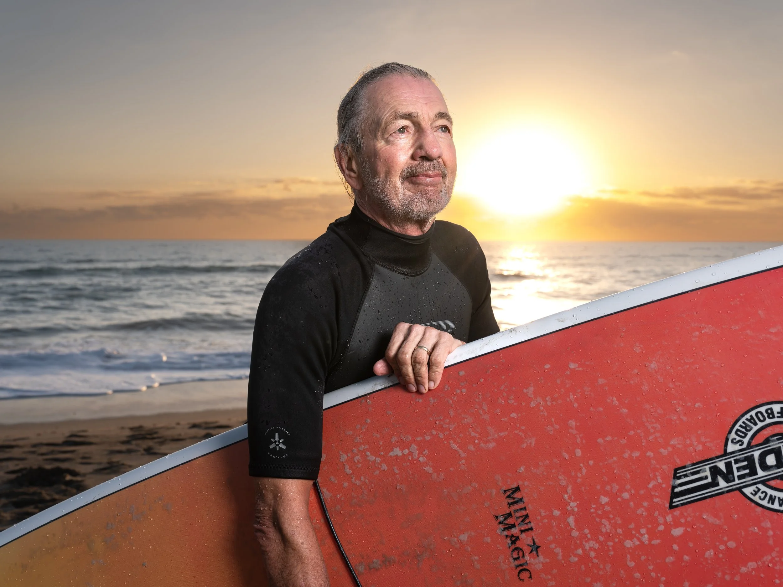 An older man holding a red surfboard on a beach at sunset, wearing a black wetsuit.