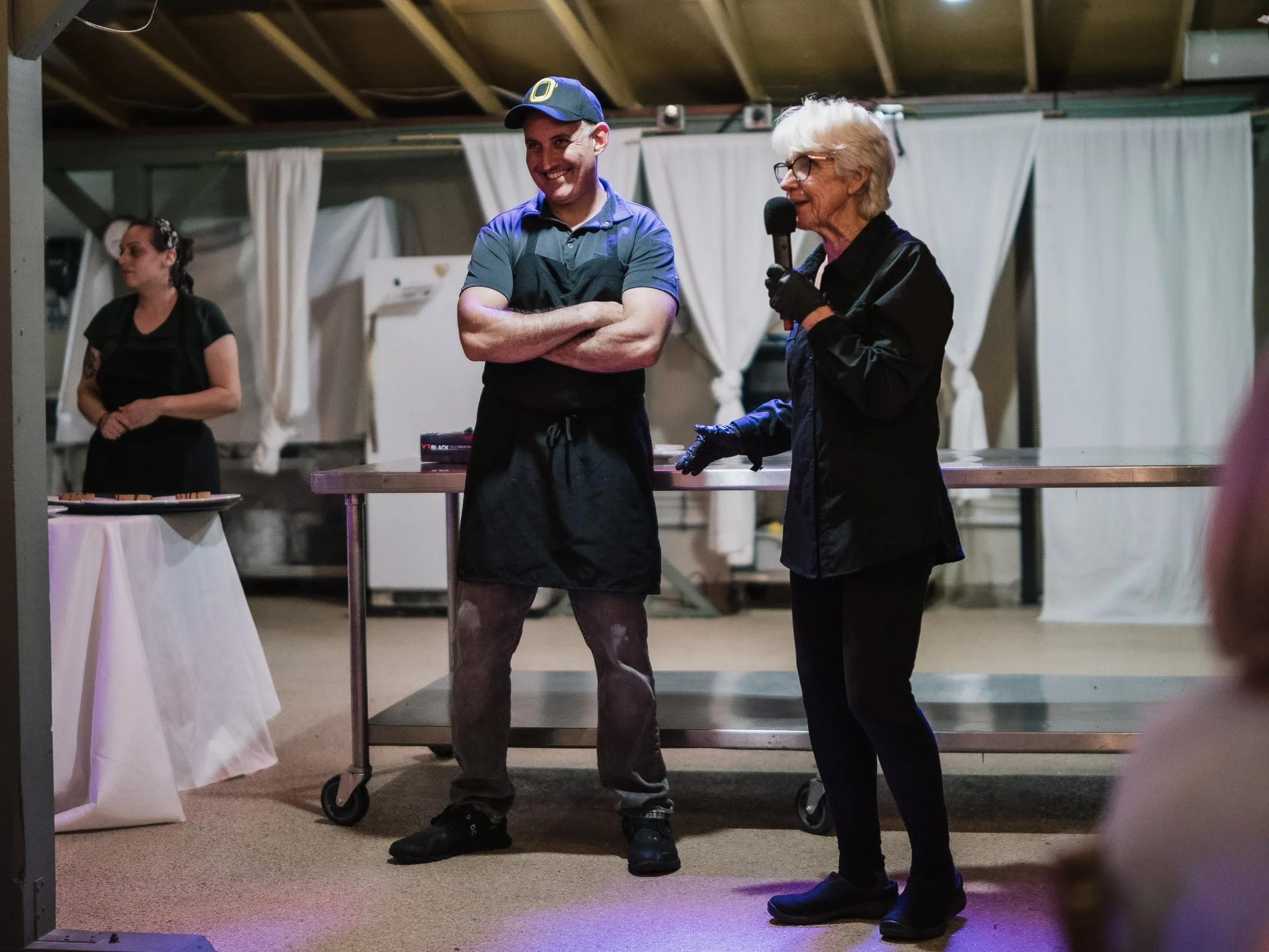 A woman with white hair, glasses, and black gloves holding a microphone, talking to a man in a gray shirt, black apron, and cap at an indoor event. Another woman in black in the background.