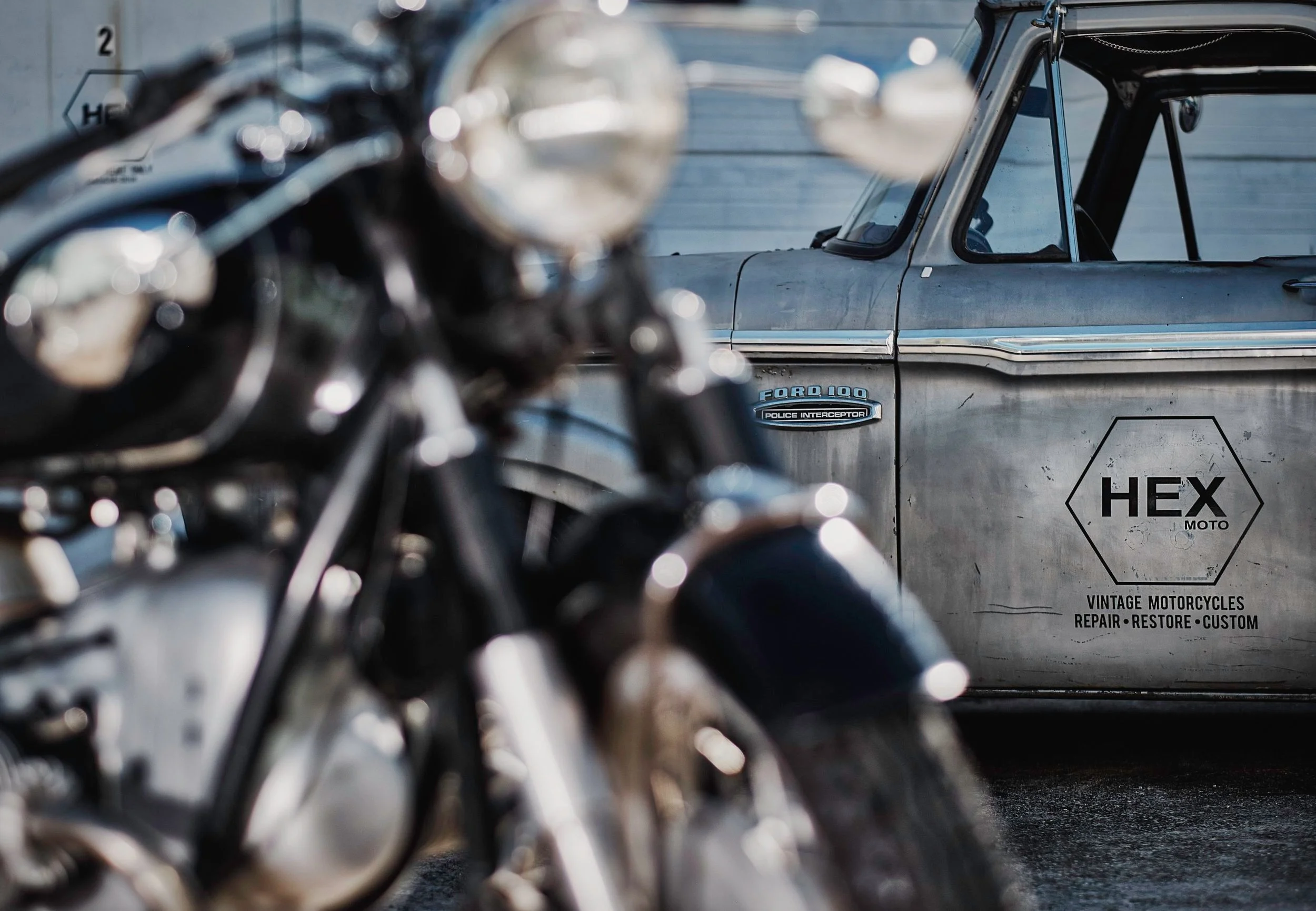 Close-up of a vintage motorcycle with shiny black and chrome parts in the foreground, and a weathered silver vintage police car with a Hex Moto logo and 
