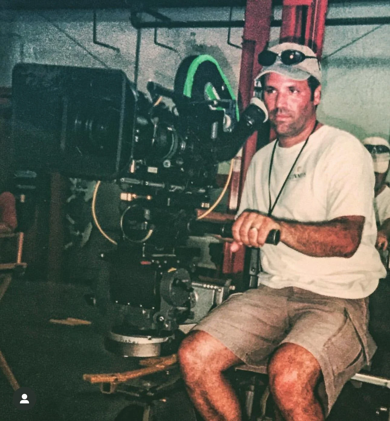A man sitting outdoors with a professional video camera on a tripod, wearing a white t-shirt, khaki shorts, and a baseball cap with sunglasses on top. He appears to be a filmmaker or cinematographer.