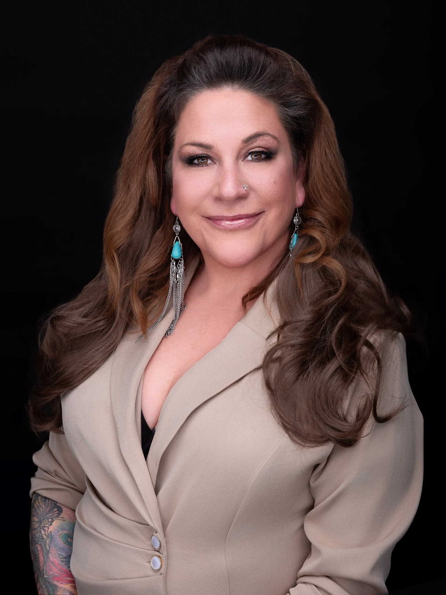 A woman with long wavy brown hair, wearing a beige blazer with rolled-up sleeves, turquoise earrings with silver chains, and a nose piercing, smiling against a black background.