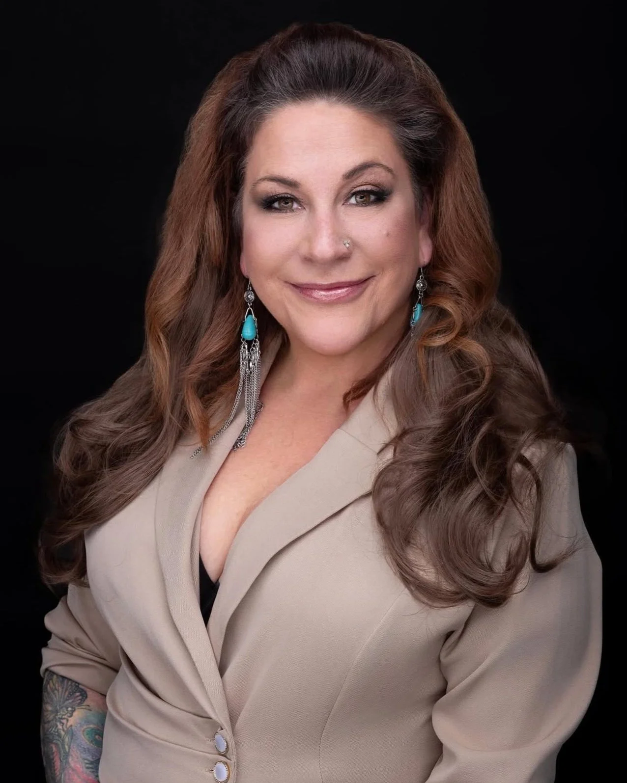 A woman with long, wavy brown hair wearing a beige blazer and turquoise earrings, smiling against a black background.
