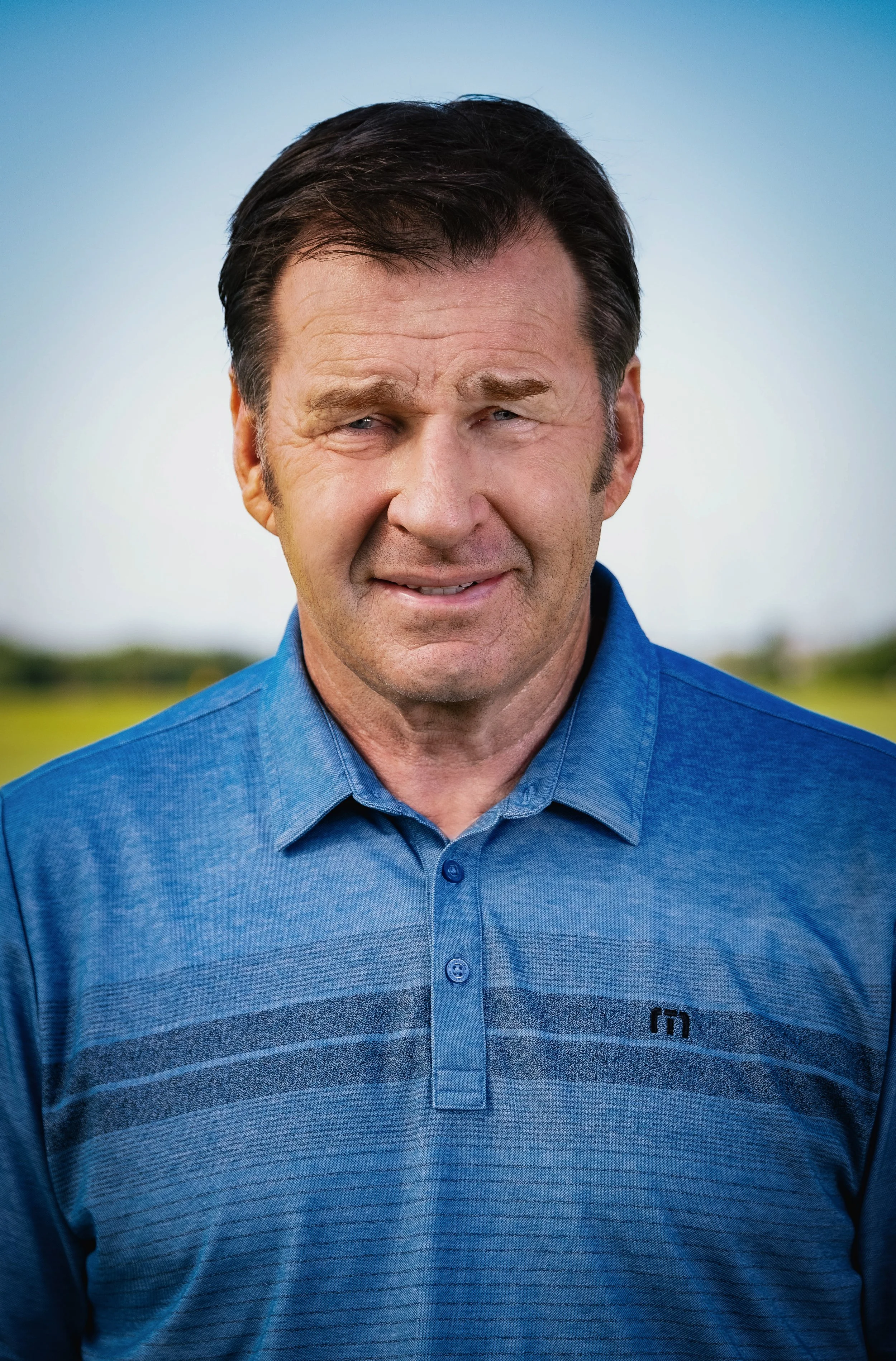 A middle-aged man with dark hair, wearing a blue collared shirt, outdoors with a blurred green landscape background, making a grimacing facial expression.