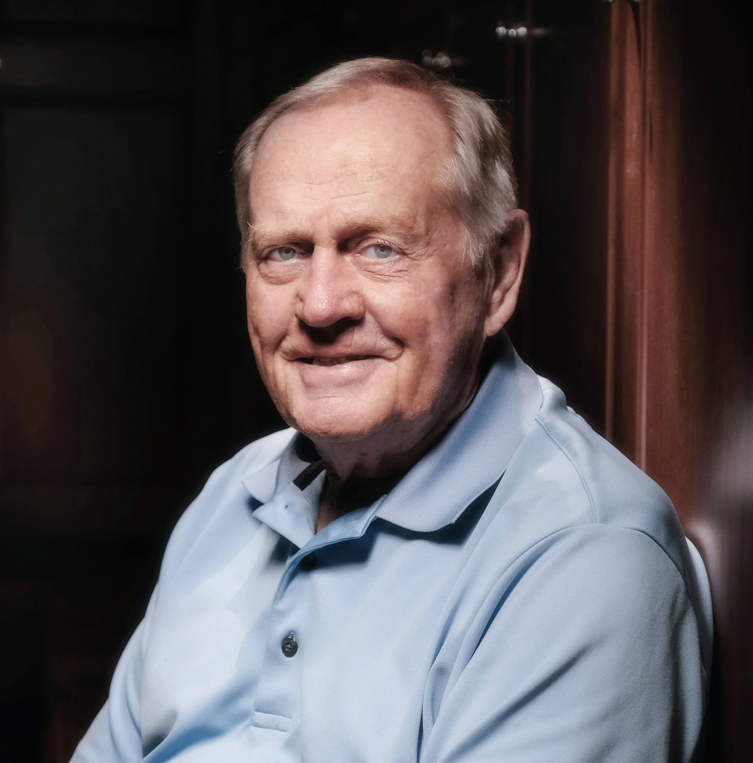 Older man with light hair and blue eyes, wearing a light blue collared shirt, smiling and leaning against a wooden wall.