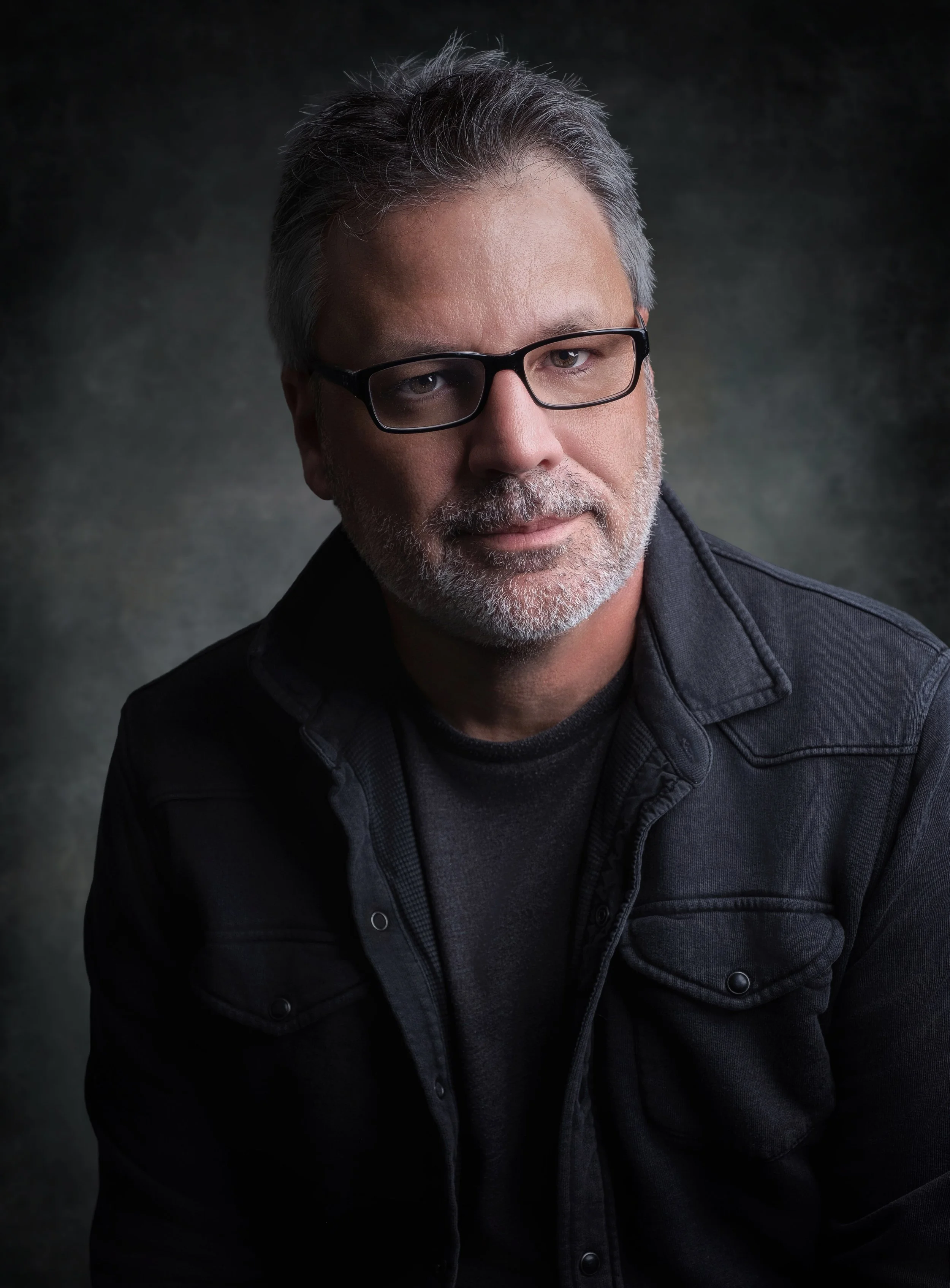 A middle-aged man with gray hair, beard, and glasses, wearing a black jacket, looking at the camera with a neutral expression.