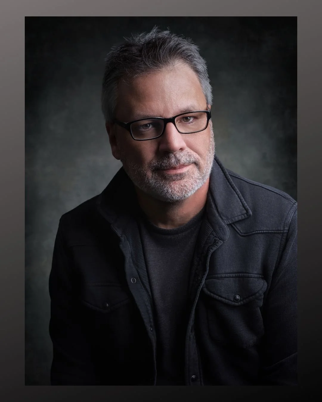 Paul Piasecki, wearing glasses and a dark jacket, looks at the camera against a dark, textured background.