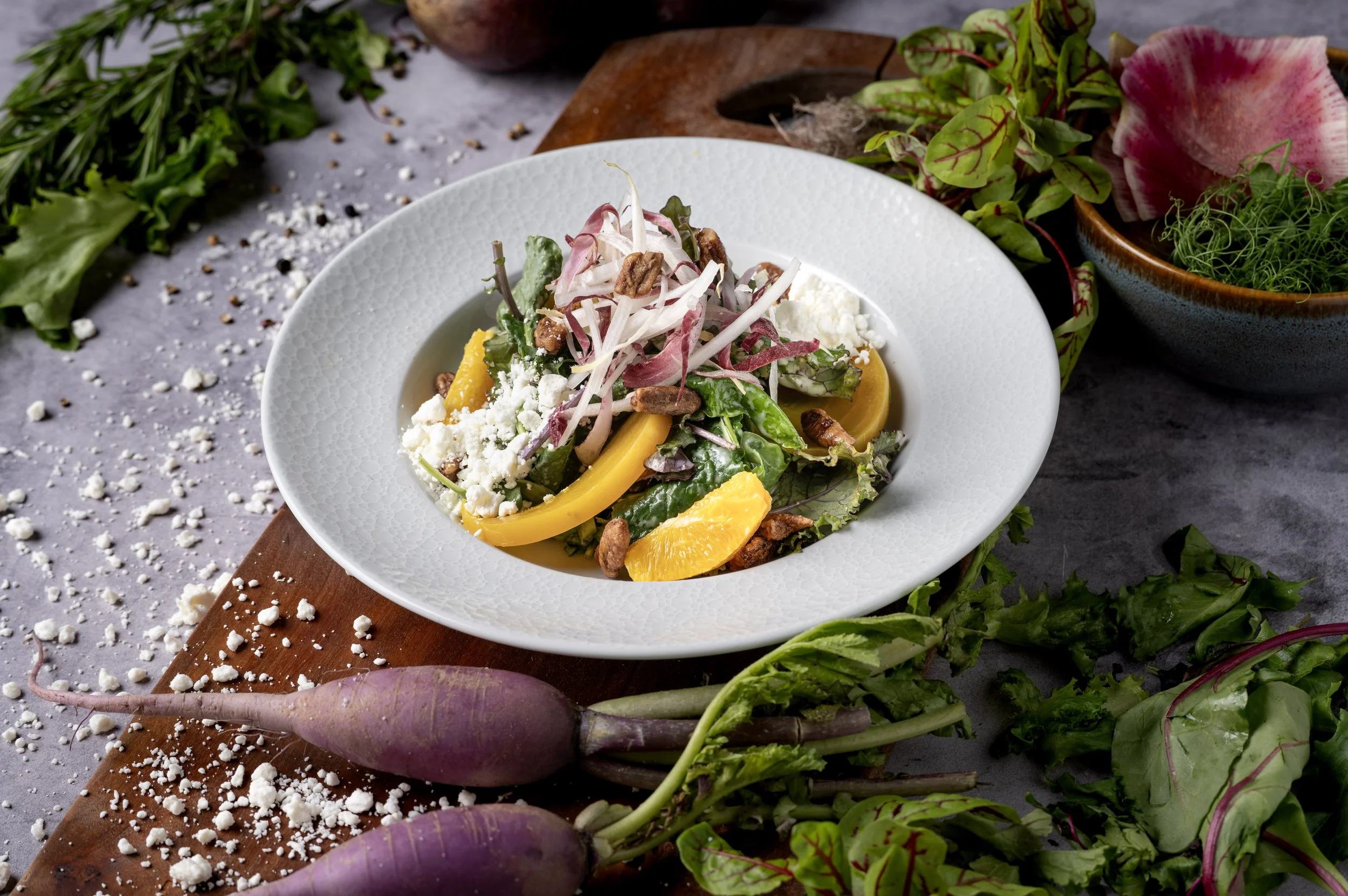 A fresh salad with mixed greens, yellow bell peppers, crumbled cheese, and nuts in a white bowl, on a table surrounded by fresh beets, herbs, and scattered cheese.