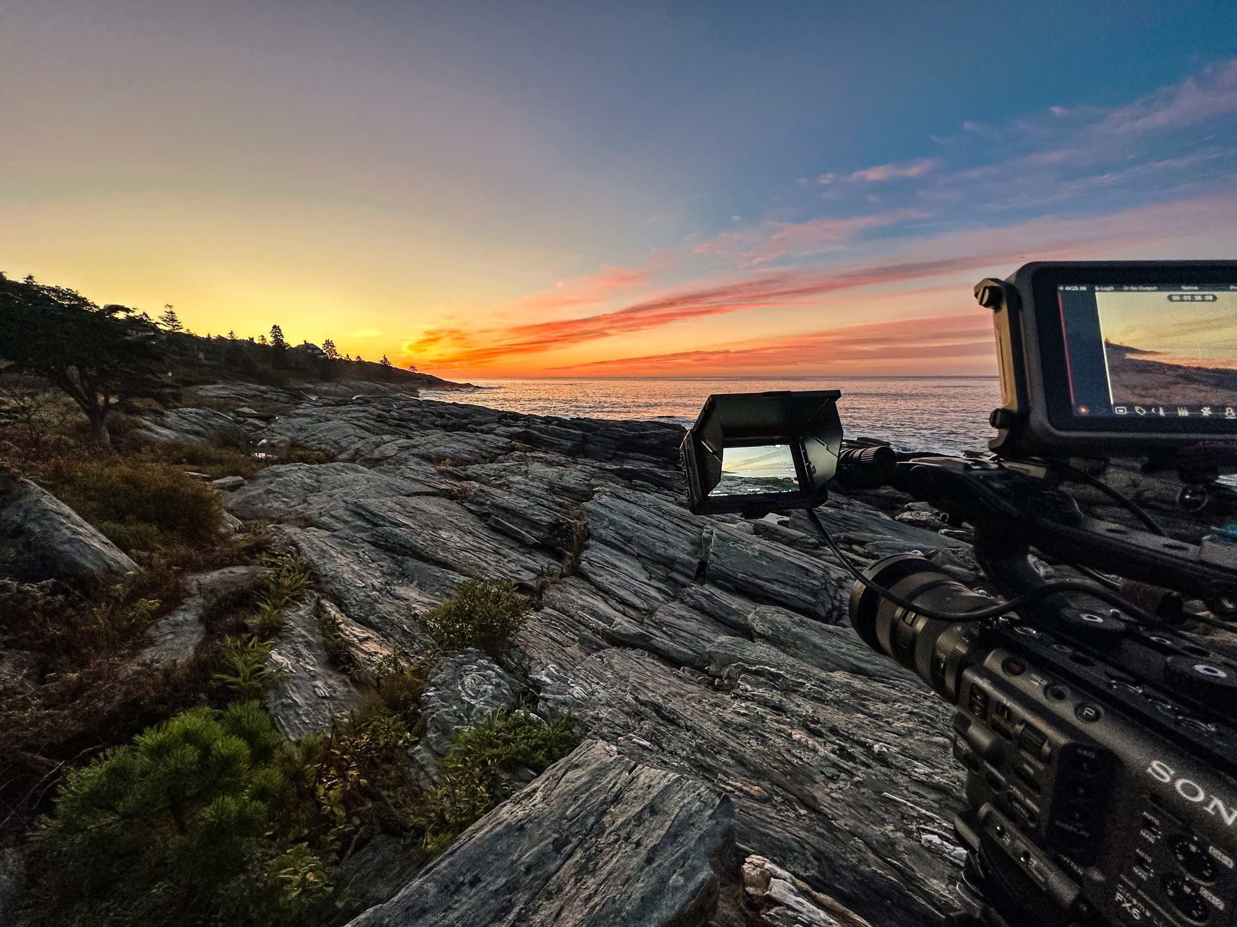 A camera set up on rocky coastline capturing sunset over the ocean with colorful clouds and silhouetted trees in the distance.