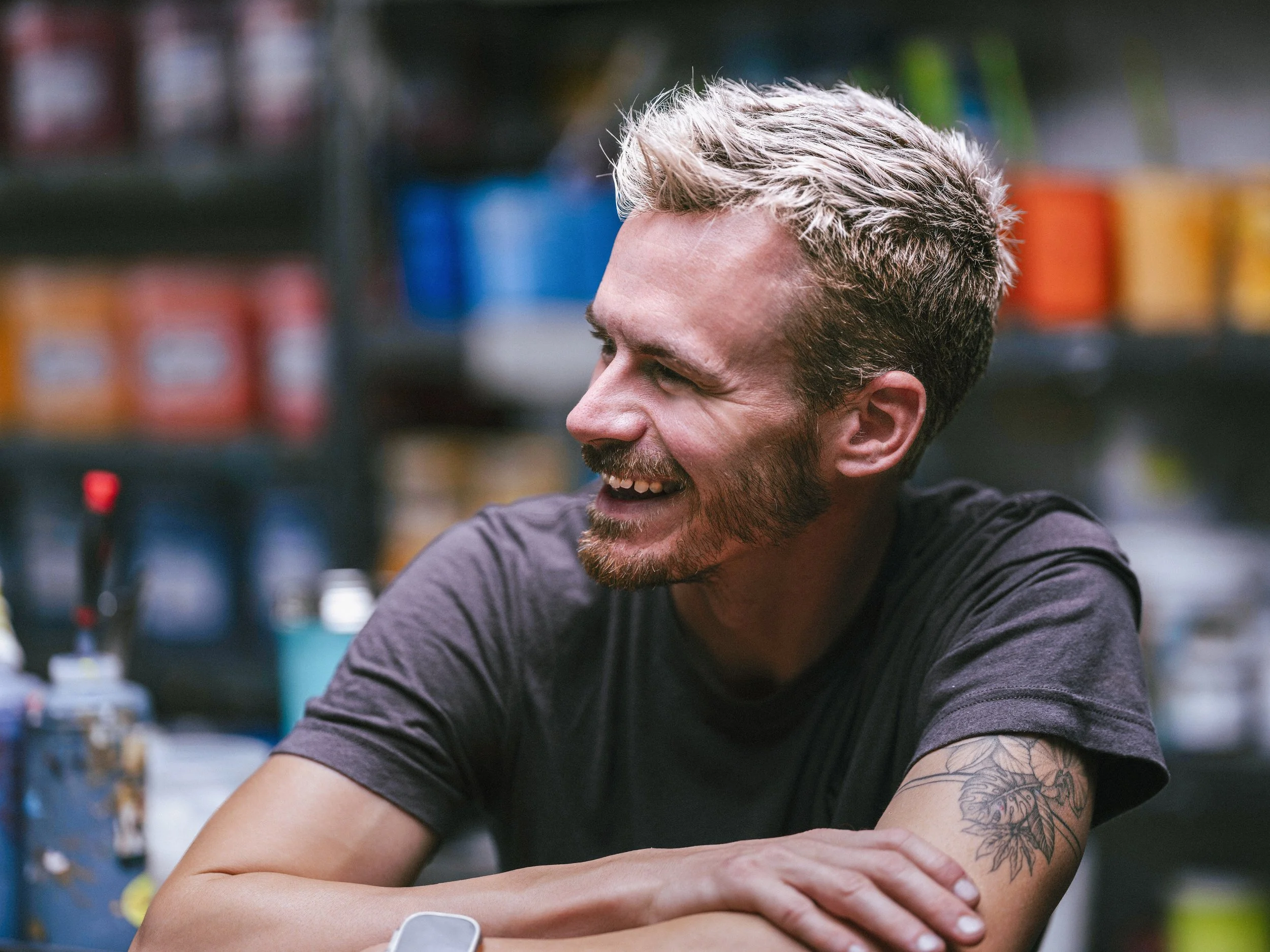A man with blond hair, a beard, and a tattoo on his arm, smiling and sitting at a table in a store with shelves of colorful products in the background.