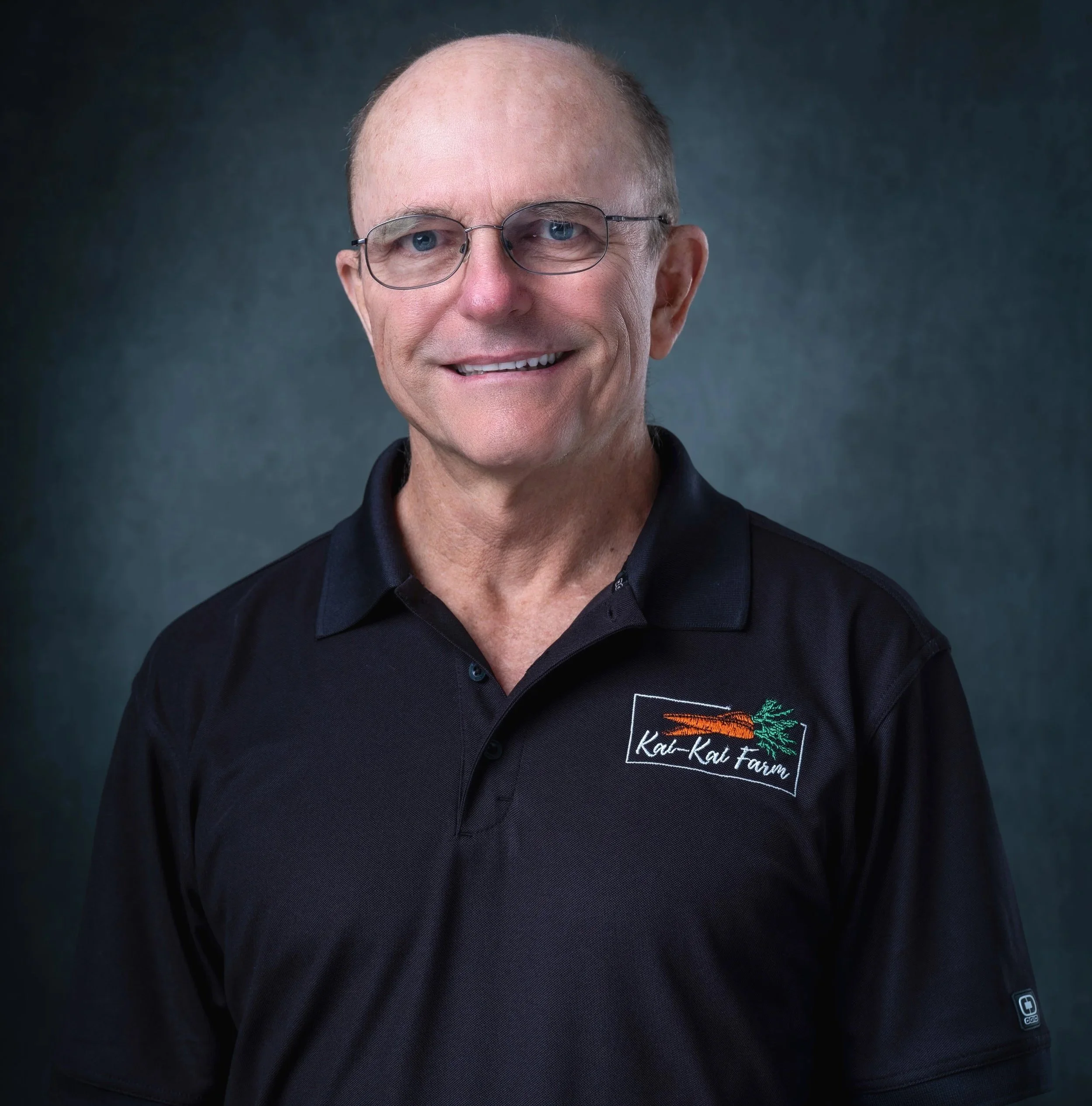 Close-up portrait of a middle-aged man with short hair, glasses, and a smile, wearing a black polo shirt with a logo that reads 'Kau Kau Farm' and a carrot graphic, against a dark, blurred background.