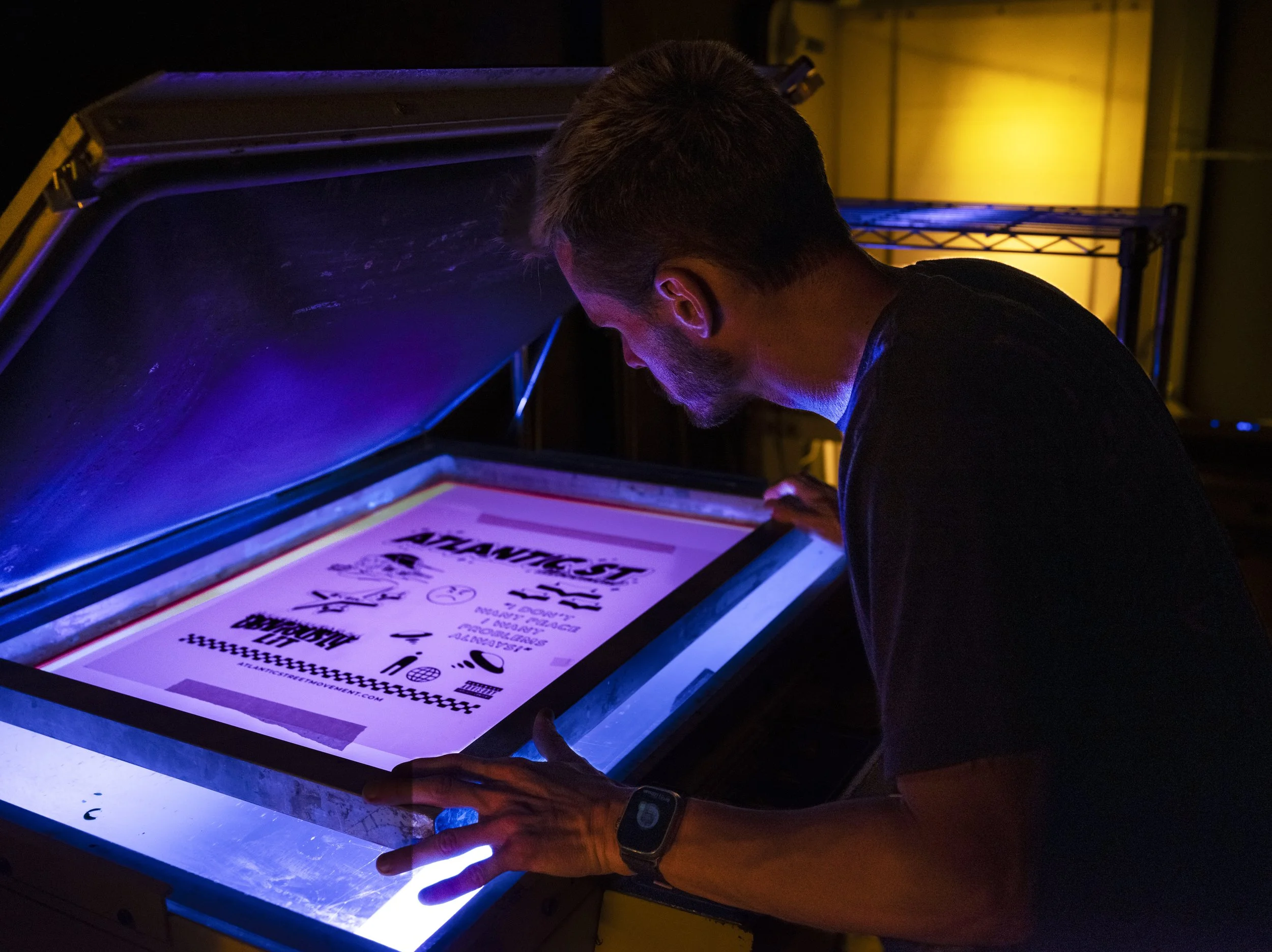 A man looking at a lightbox displaying printed flyers or posters with black text and graphics in a dark room with colored lighting.