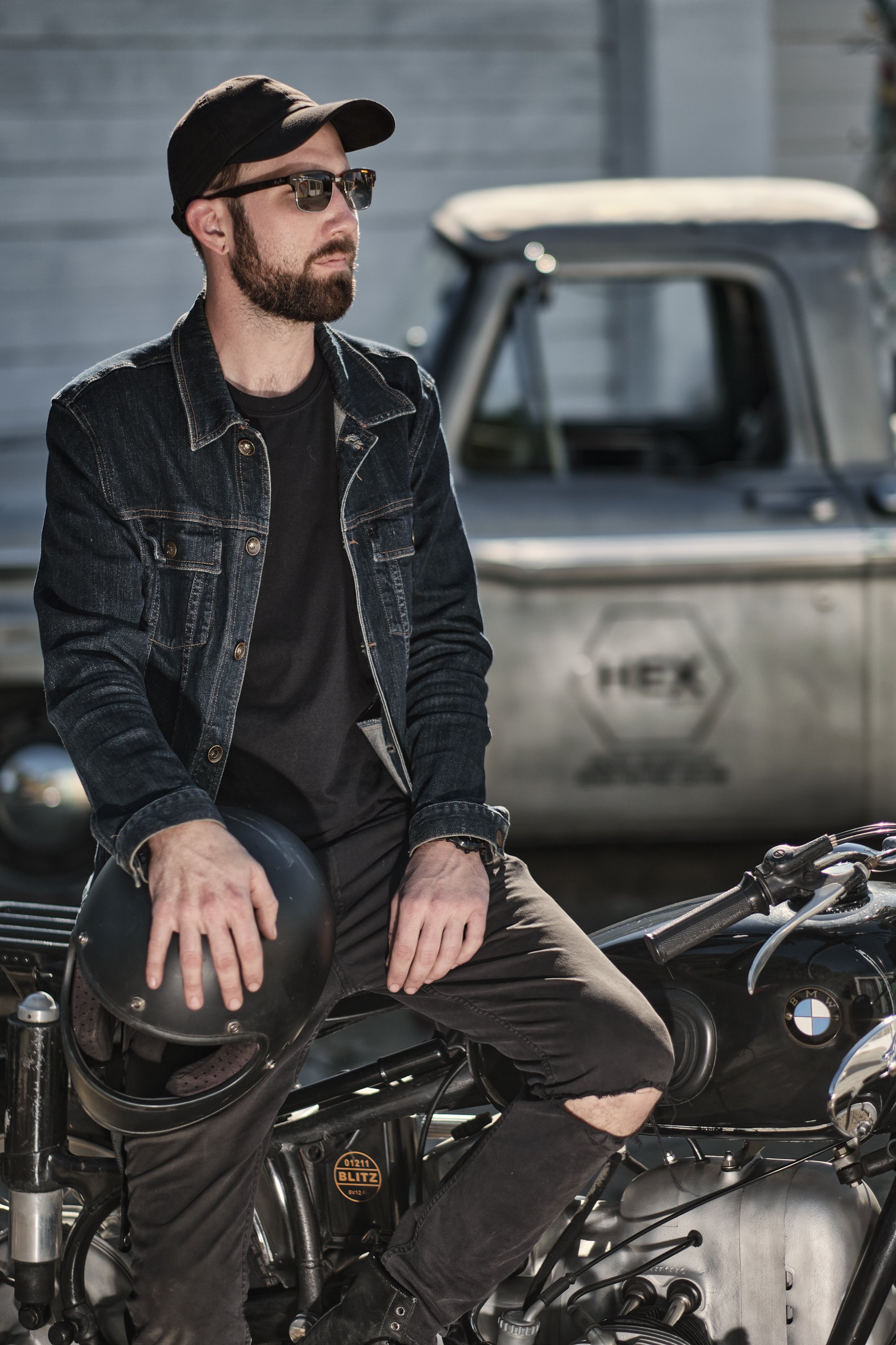 A man with sunglasses, a hat, and a denim jacket sitting on a motorcycle, holding a helmet, with a vintage truck in the background.
