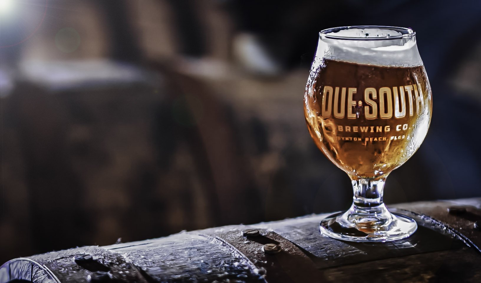 A glass of beer with the logo 'DUE SOUTH BREWING CO' on it, sitting on a wooden surface with blurred background.