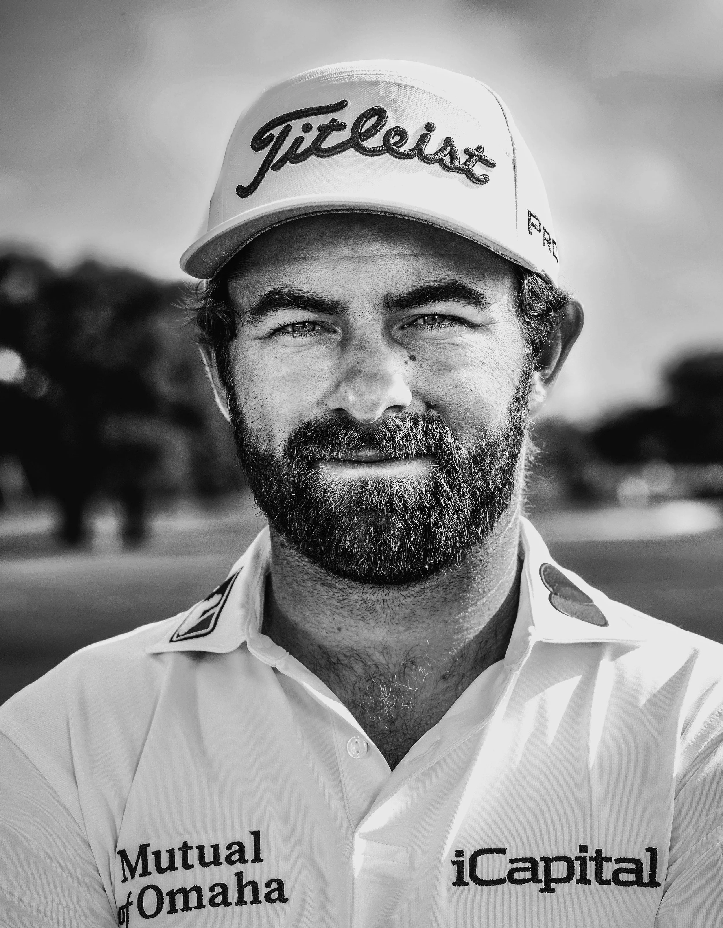 Black and white portrait of a man with a beard wearing a white golf shirt with sponsor logos and a white cap with 'Titleist' written on it, outdoors with blurred trees in the background.