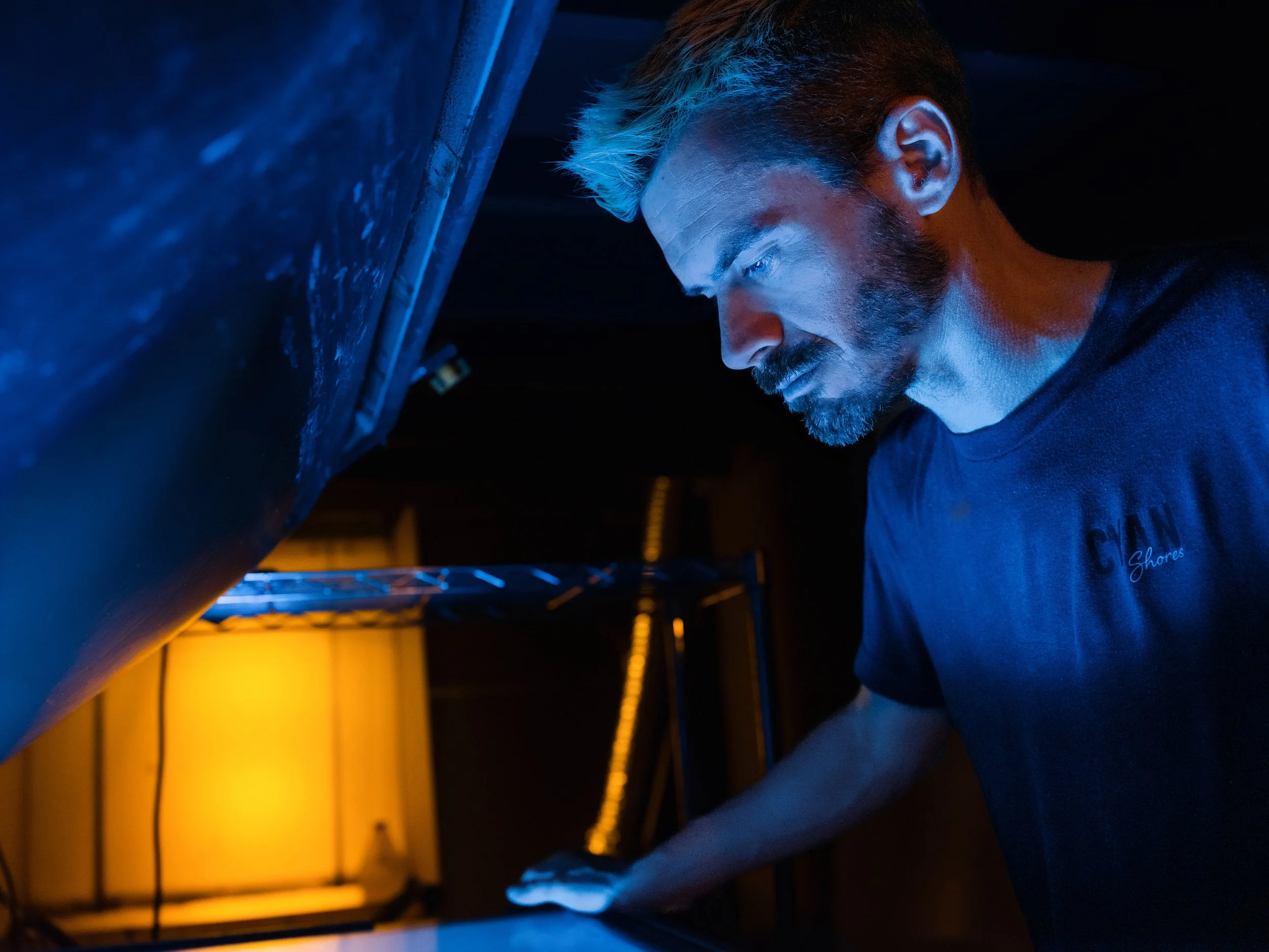 A man with blonde hair and a beard looks down at a surface, illuminated by blue and orange lighting, in a dark environment.