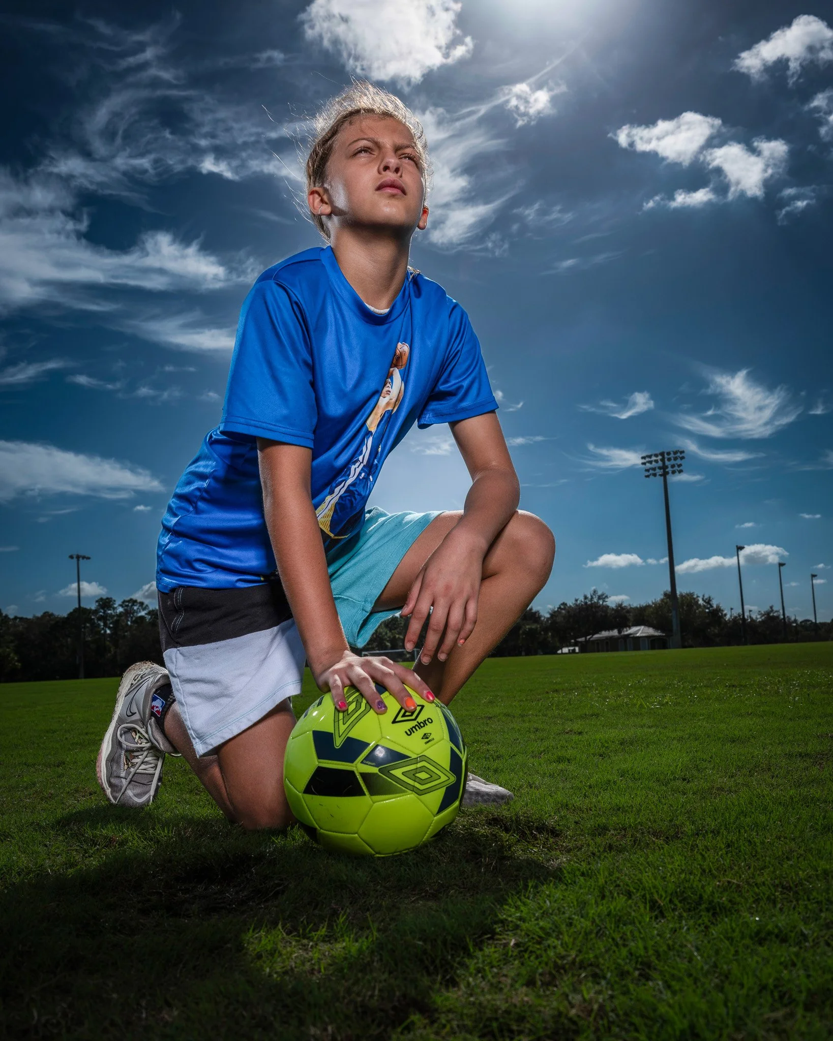 A young girl in a blue sports jersey kneeling on grass football field, holding a bright yellow soccer ball, under a partly cloudy sky.