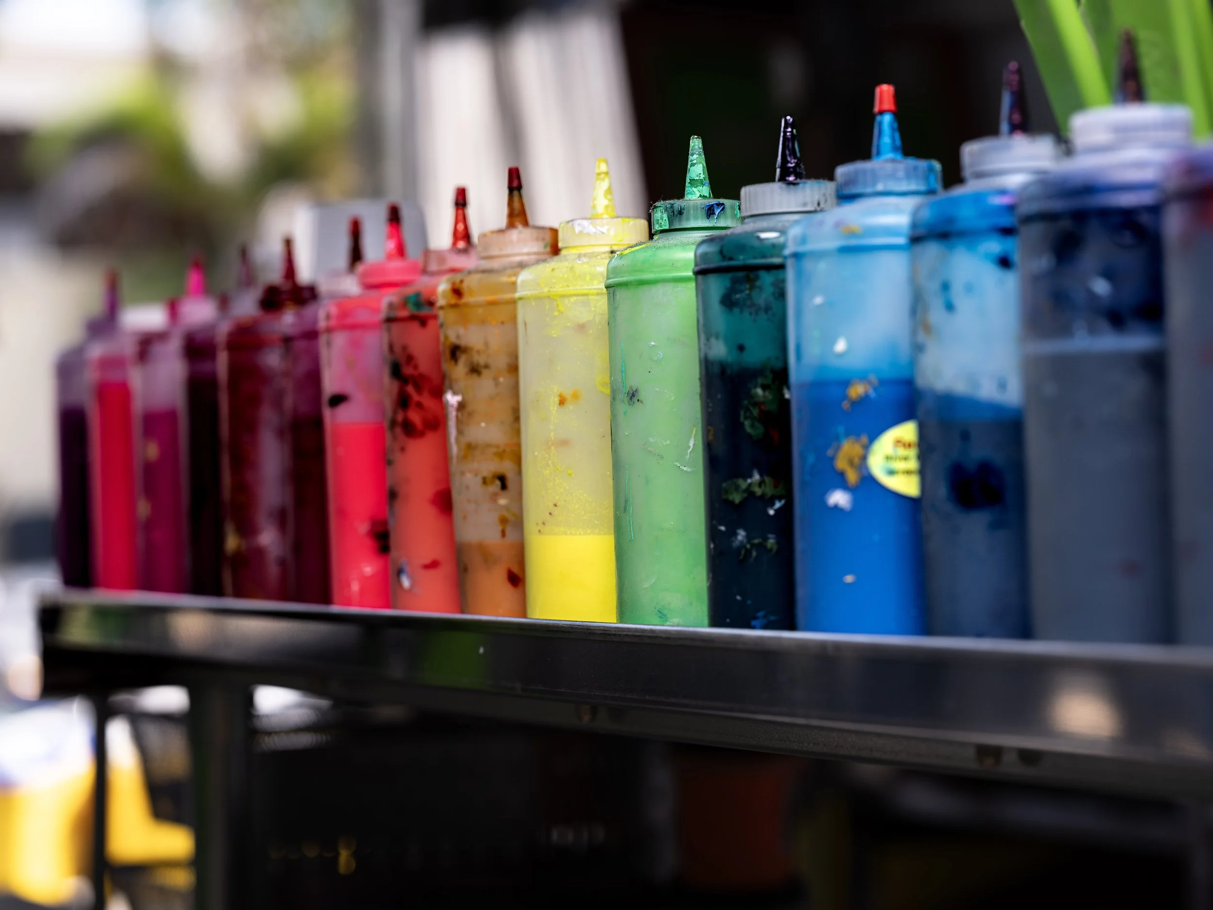 Various bottles of colorful acrylic paint lined up on a shelf outdoors.