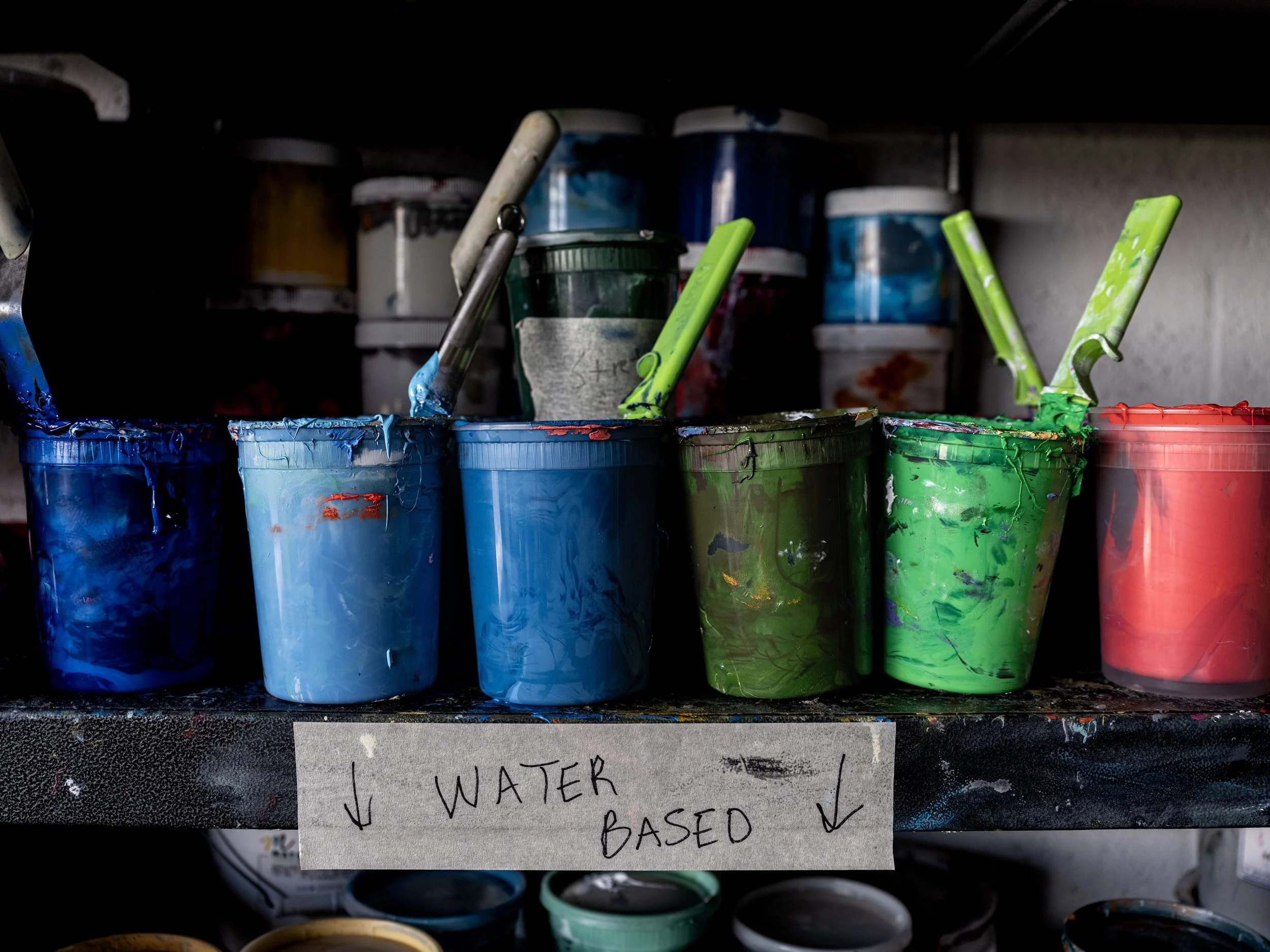 Buckets of water-based paint in blue, green, and red, with paintbrushes submerged, on a shelf labeled 'Water Based'.