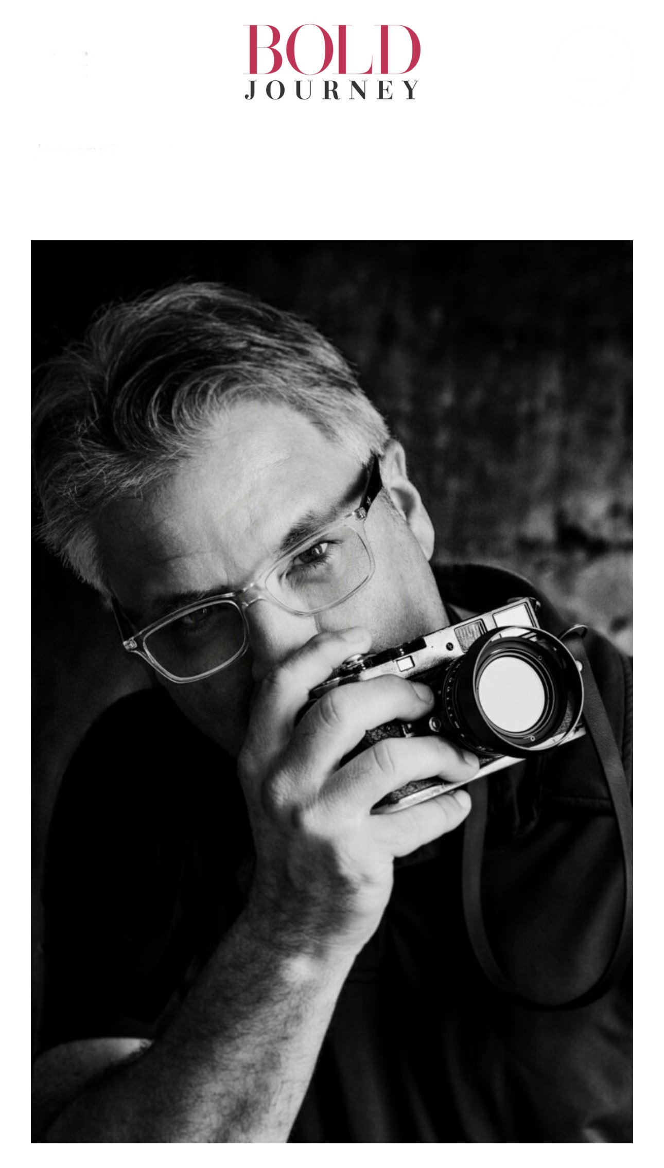 Black and white photo of a man with glasses holding a vintage camera up to his eye, looking into the lens.
