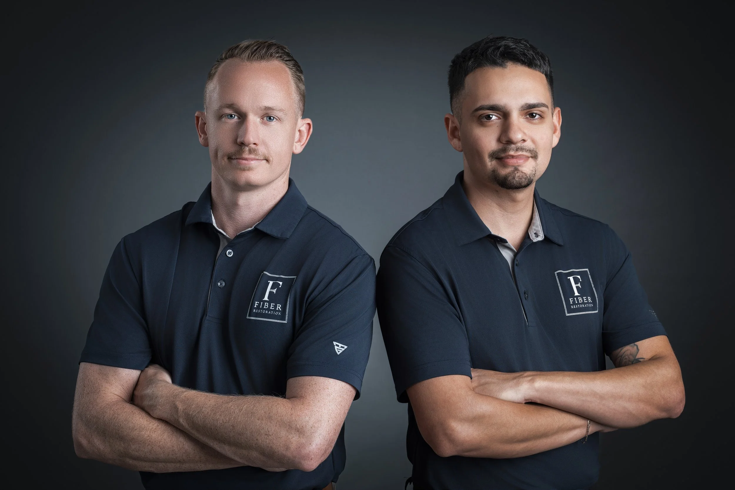 Two men standing with arms crossed, wearing navy polo shirts with a logo that says 'Fiber Restoration' on the chest, against a dark background.