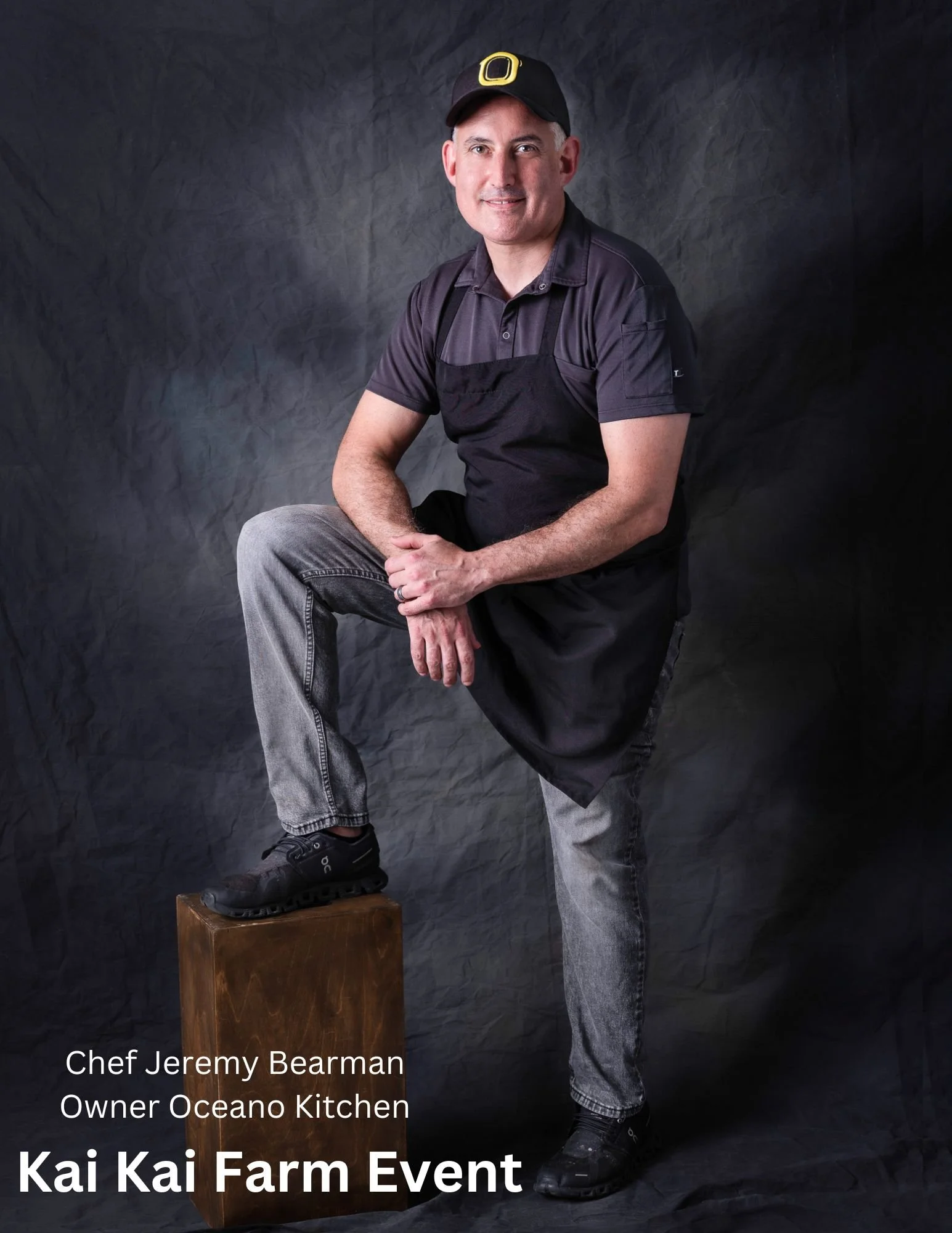 Portrait of Chef Jeremy Bearman, owner of Oceano Kitchen, standing with one foot on a wooden block against a dark textured background, wearing a black apron, black cap with a yellow 'O' logo, black shirt, gray jeans, and black sneakers, at the Kai Ka