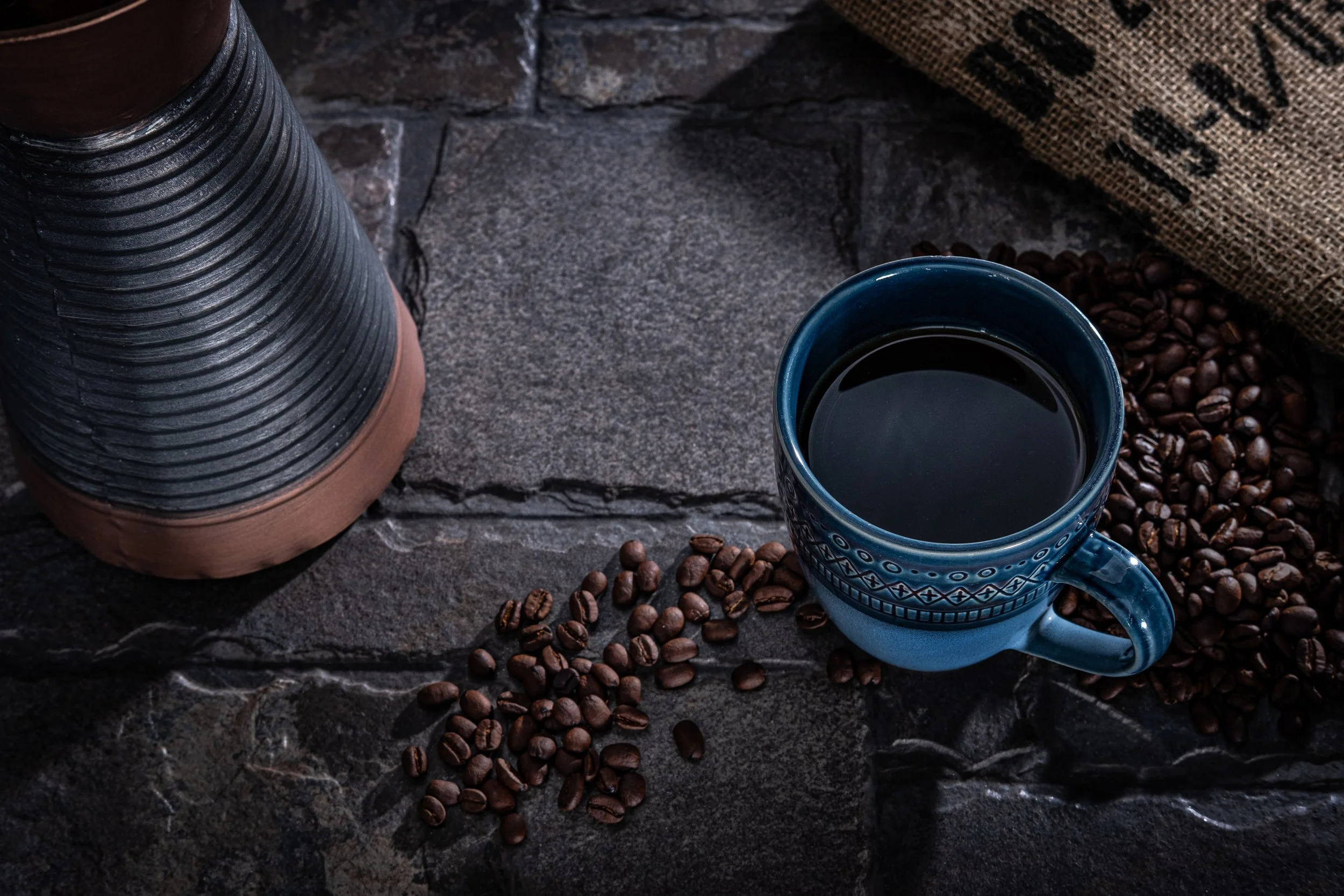 A cup of black coffee on a dark stone surface surrounded by scattered coffee beans, with a coffee grinder to the left and a burlap sack in the background.