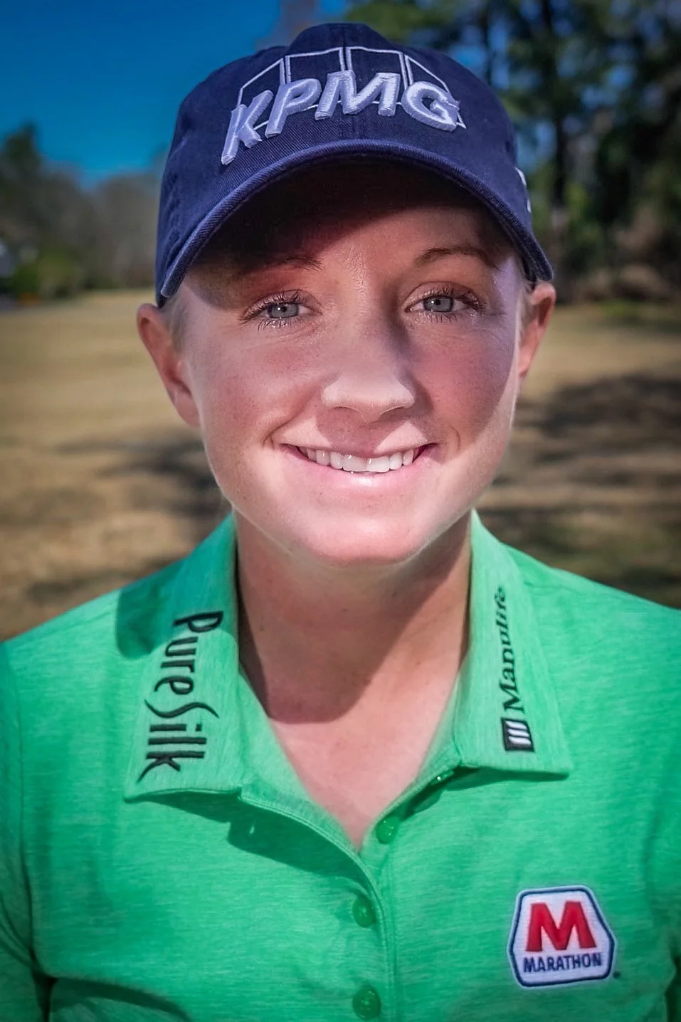 A smiling woman wearing a green polo shirt with sponsor logos and a navy baseball cap with 'KPMG' embroidered on it, outdoors with trees in the background.