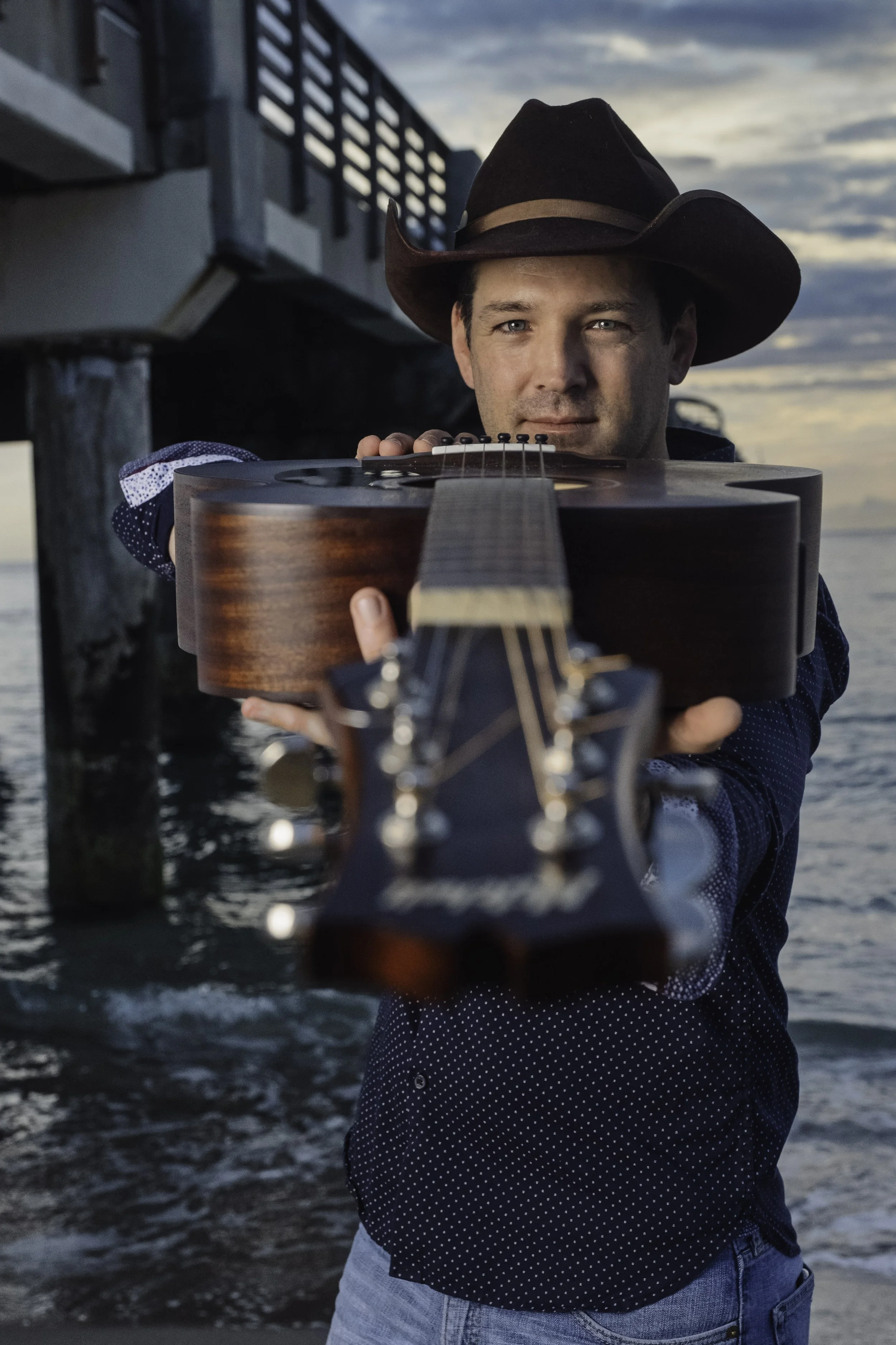 A man in a cowboy hat holding a guitar towards the camera on a beach next to a pier at sunset.