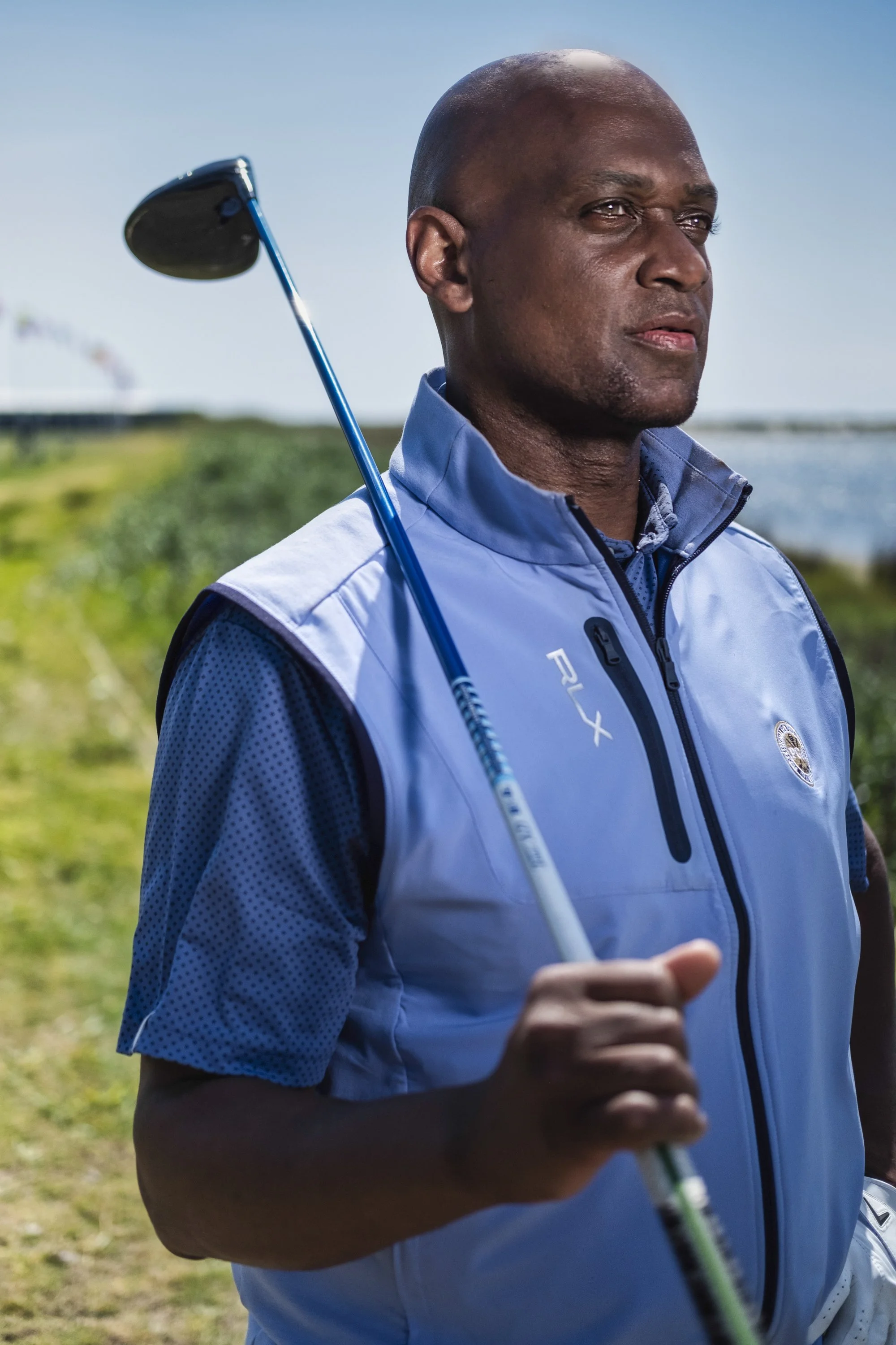 A man is standing outdoors near water, holding a golf club over his shoulder, wearing a light blue sports jacket.