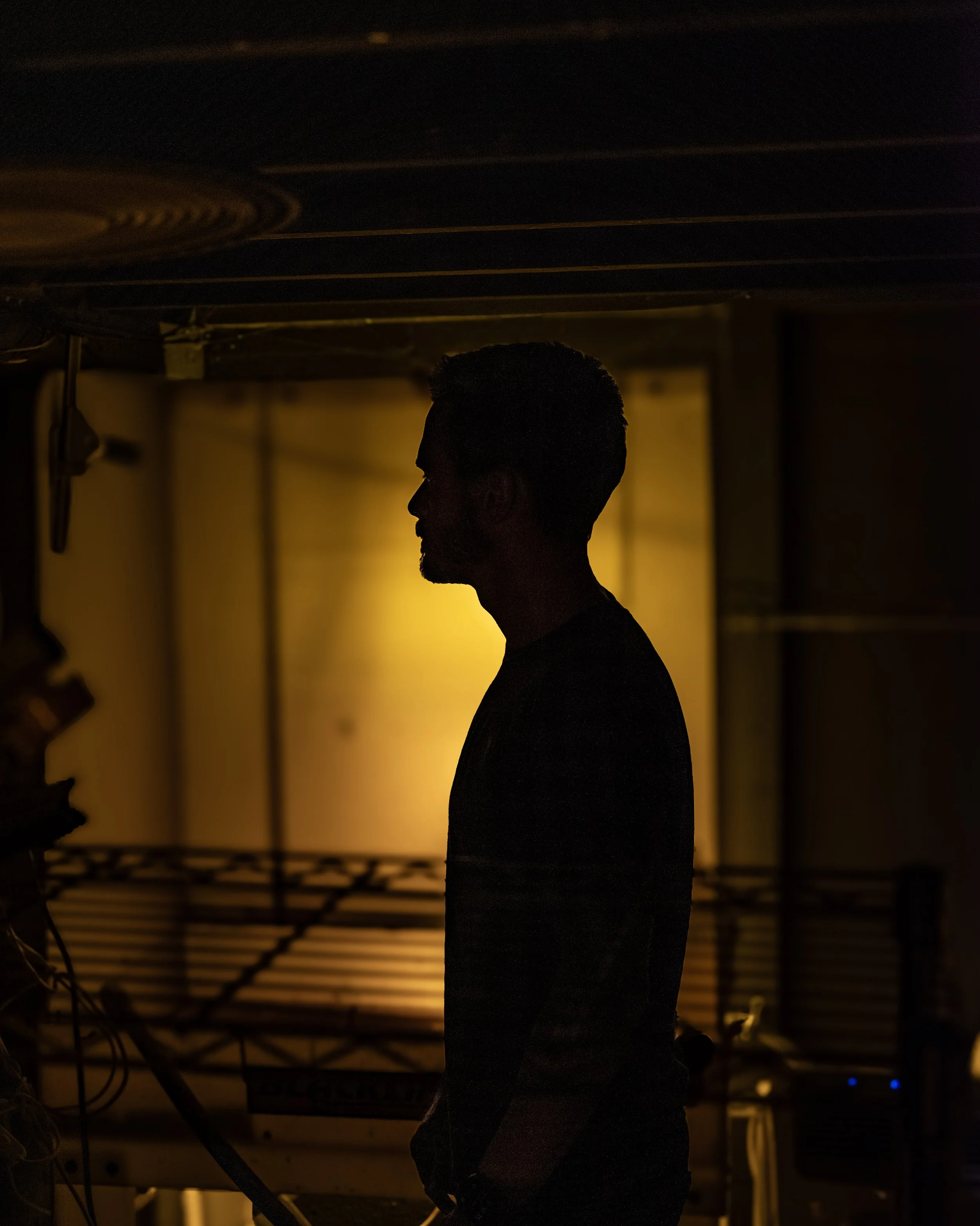 Silhouette of a man standing indoors with a warm yellow backlight, showing profile view, surrounded by equipment and dark environment.
