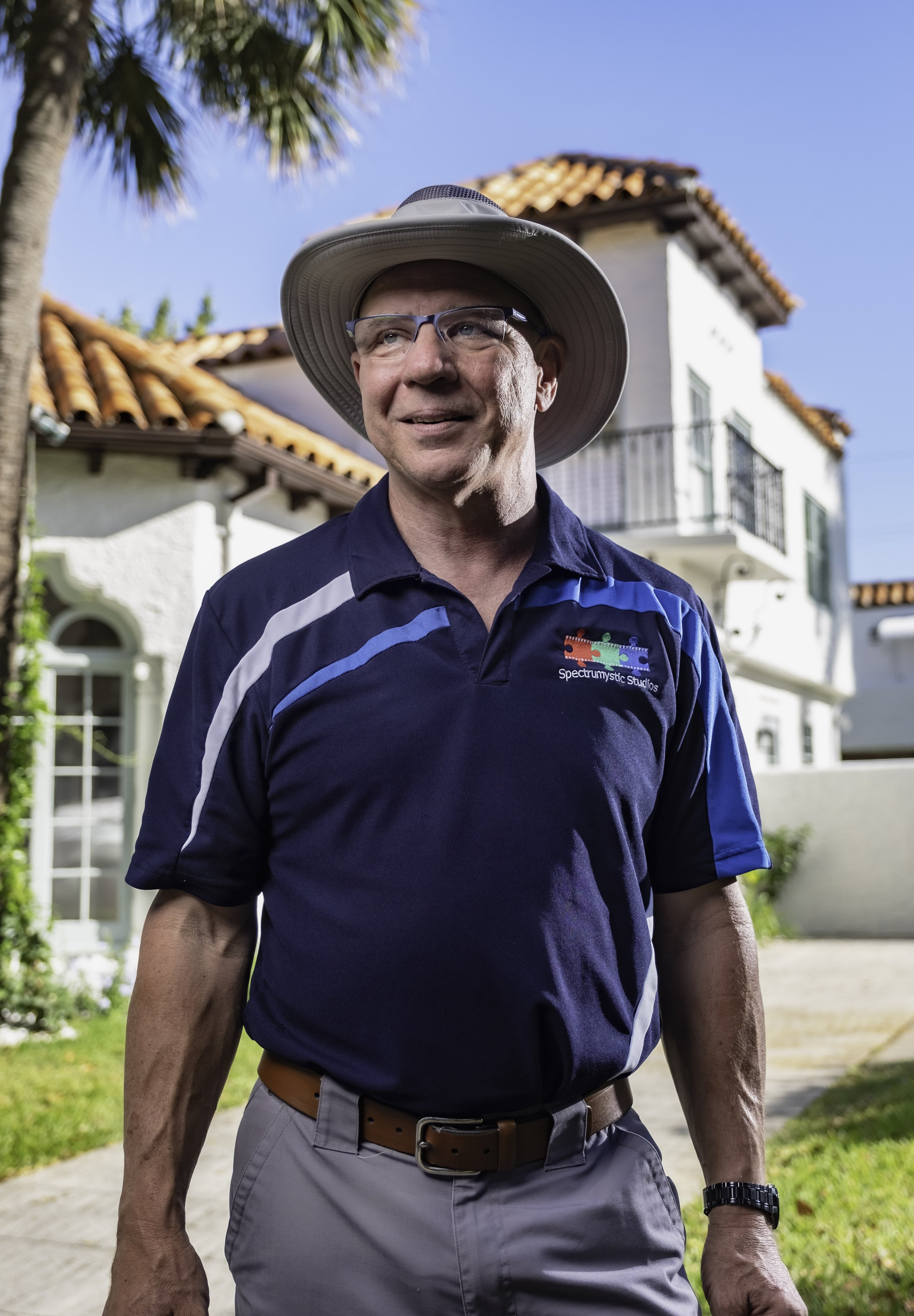 A man in a blue Spectrumistic Studios polo shirt and wide-brimmed hat standing outdoors in front of a white house with a red tile roof.