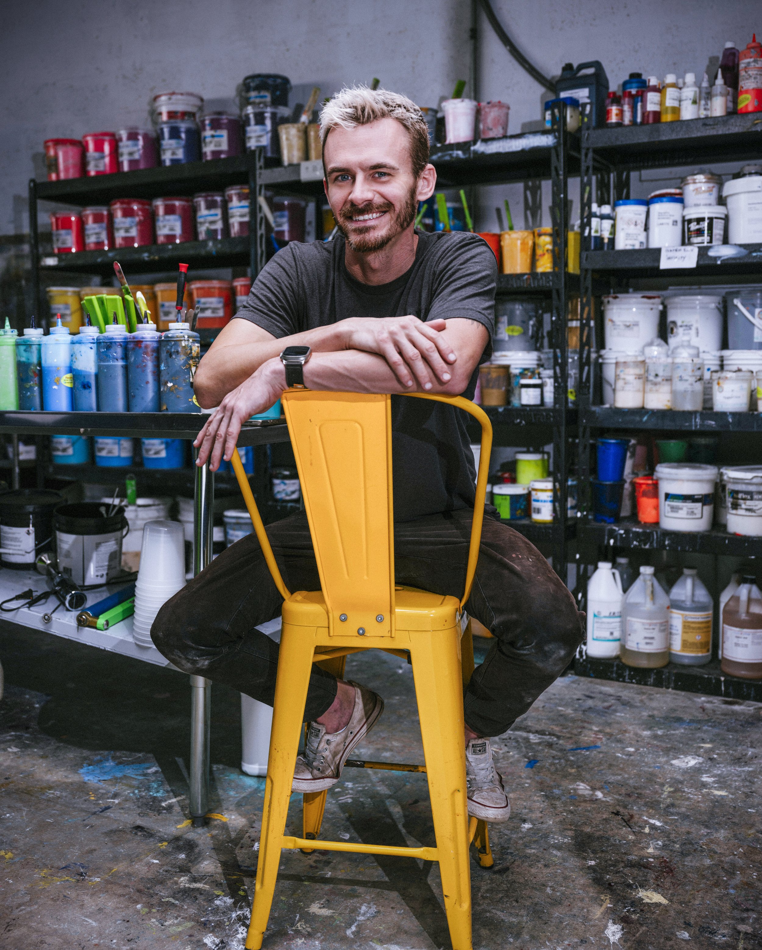 A smiling man with blond hair and a beard sitting on a yellow chair in an art studio, with shelves of paint cans and art supplies in the background.