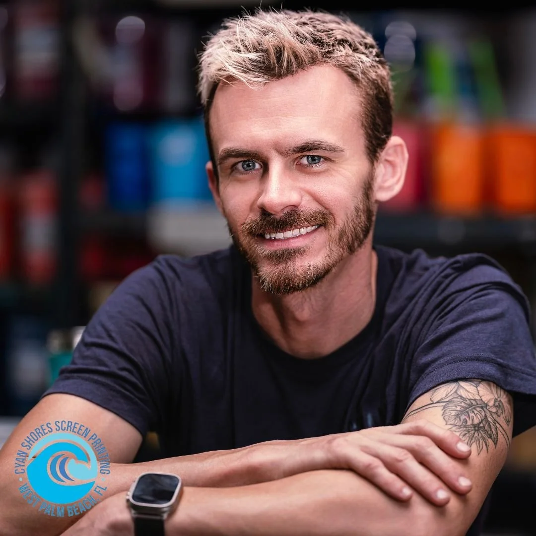 A man with a beard and blonde hair smiling at the camera, sitting at a table with arms crossed. Background features colorful cans. He is wearing a dark t-shirt and a smartwatch on his left wrist.