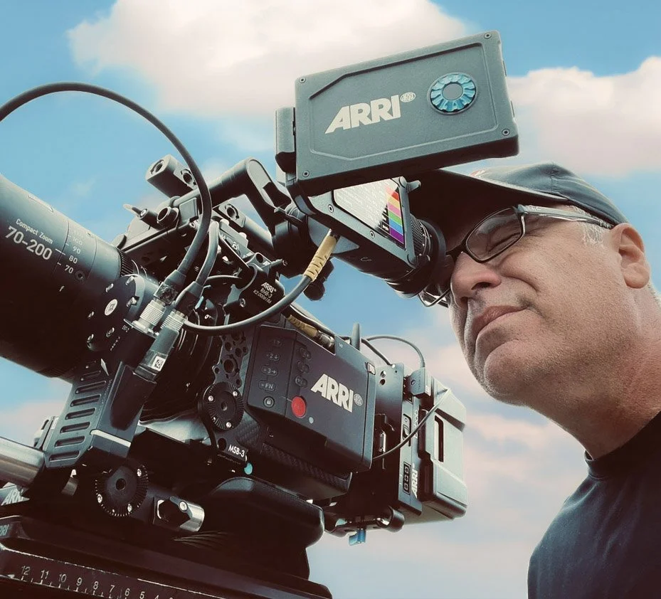 A man operating a professional ARRI digital cinema camera outdoors against a partly cloudy sky.