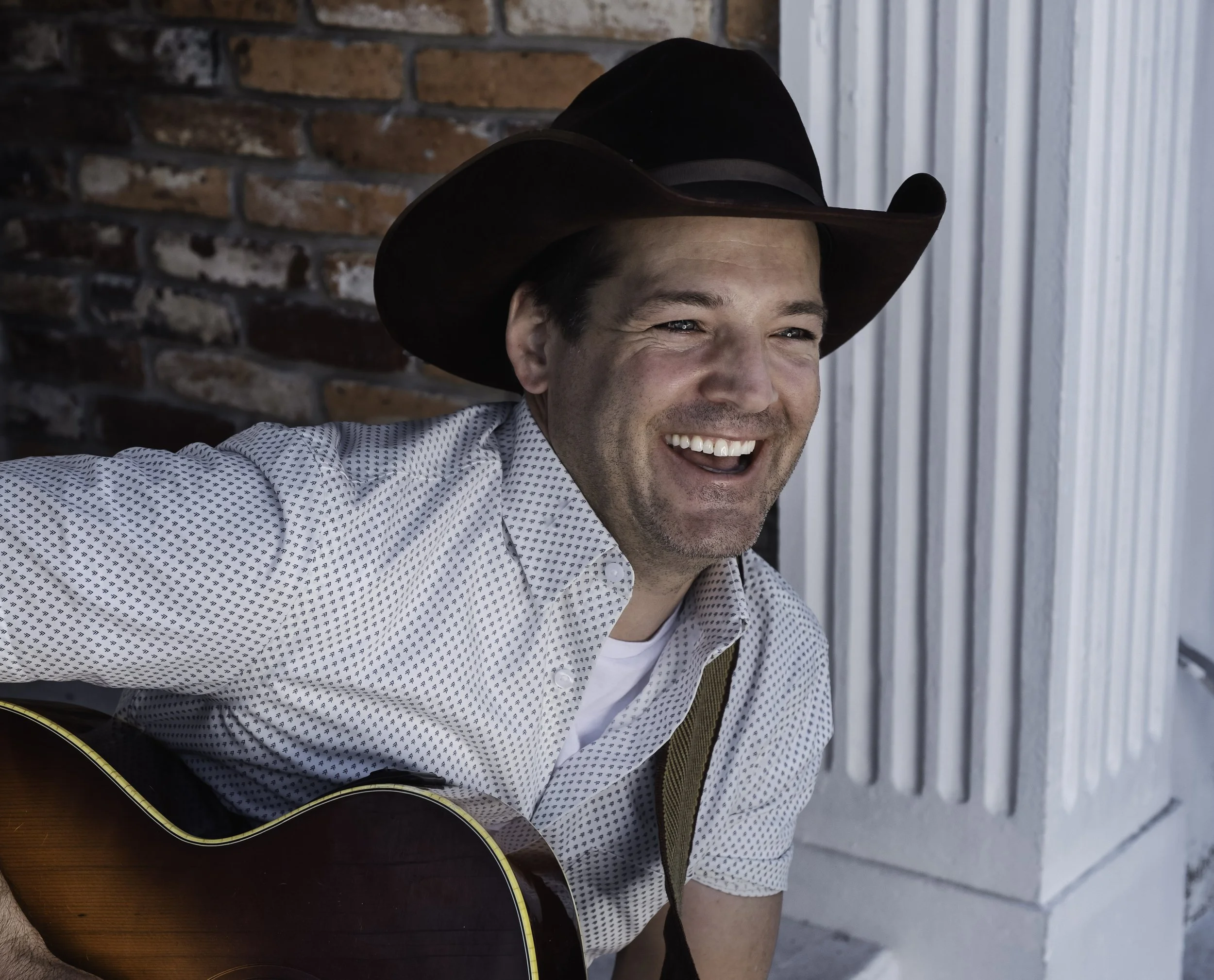 A man wearing a large dark cowboy hat is smiling while playing an acoustic guitar. He is dressed in a white shirt with small dark patterns, and there is a brick wall and metal siding behind him.