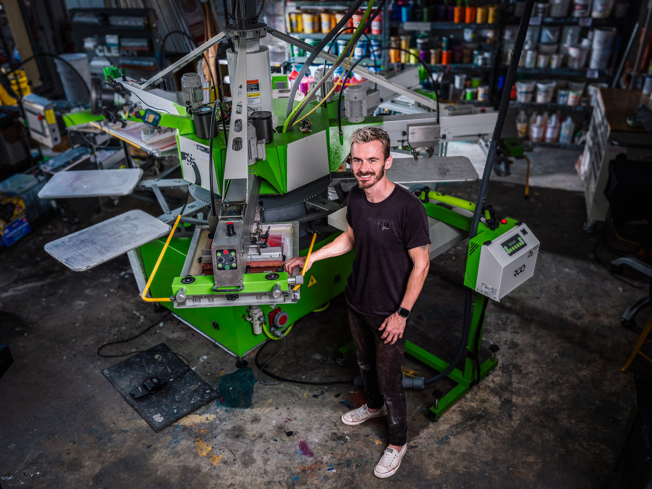 A man standing next to a large green and white industrial machine in a workshop, surrounded by tools and shelves filled with supplies.