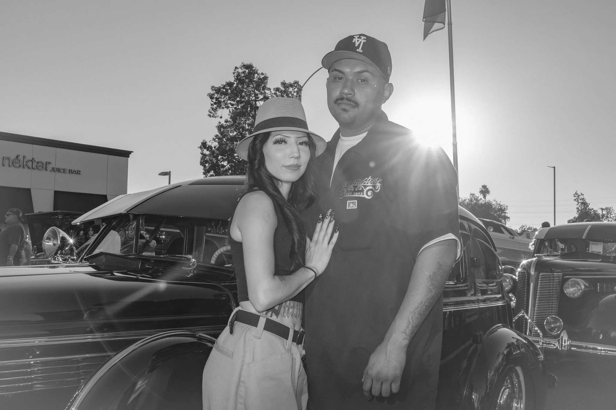 Black-and-white photo of a man and woman standing close together in front of vintage cars at an outdoor event during sunset. The woman is wearing a hat and tank top, and the man is wearing a baseball cap and a shirt.