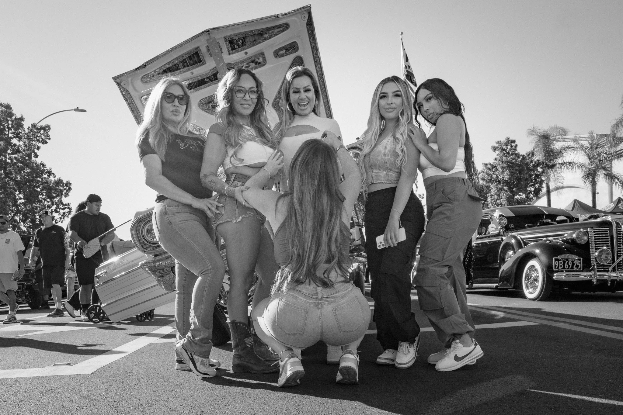 A group of seven women posing for a photo at a classic car show, with vintage cars and trees in the background.