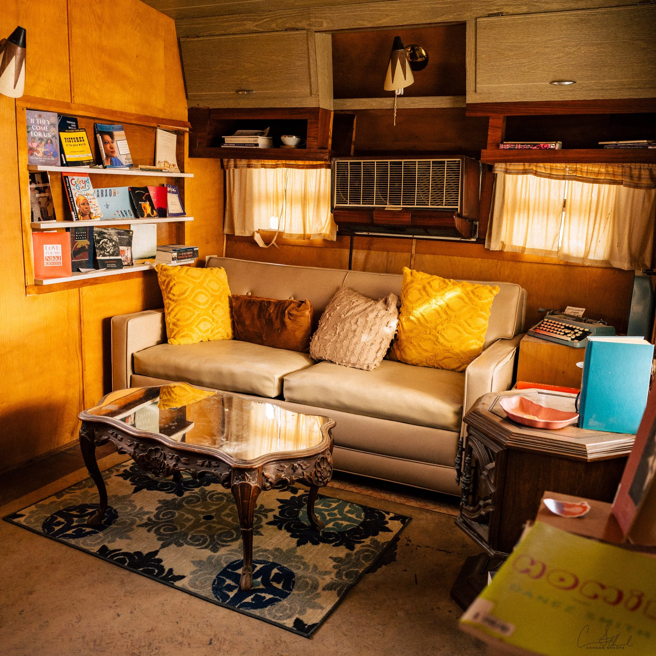 Vintage living room with a beige couch decorated with multicolored pillows, a wooden coffee table with ornate carvings, a patterned rug, books on shelves, small windows with beige curtains, and a retro air conditioning unit.