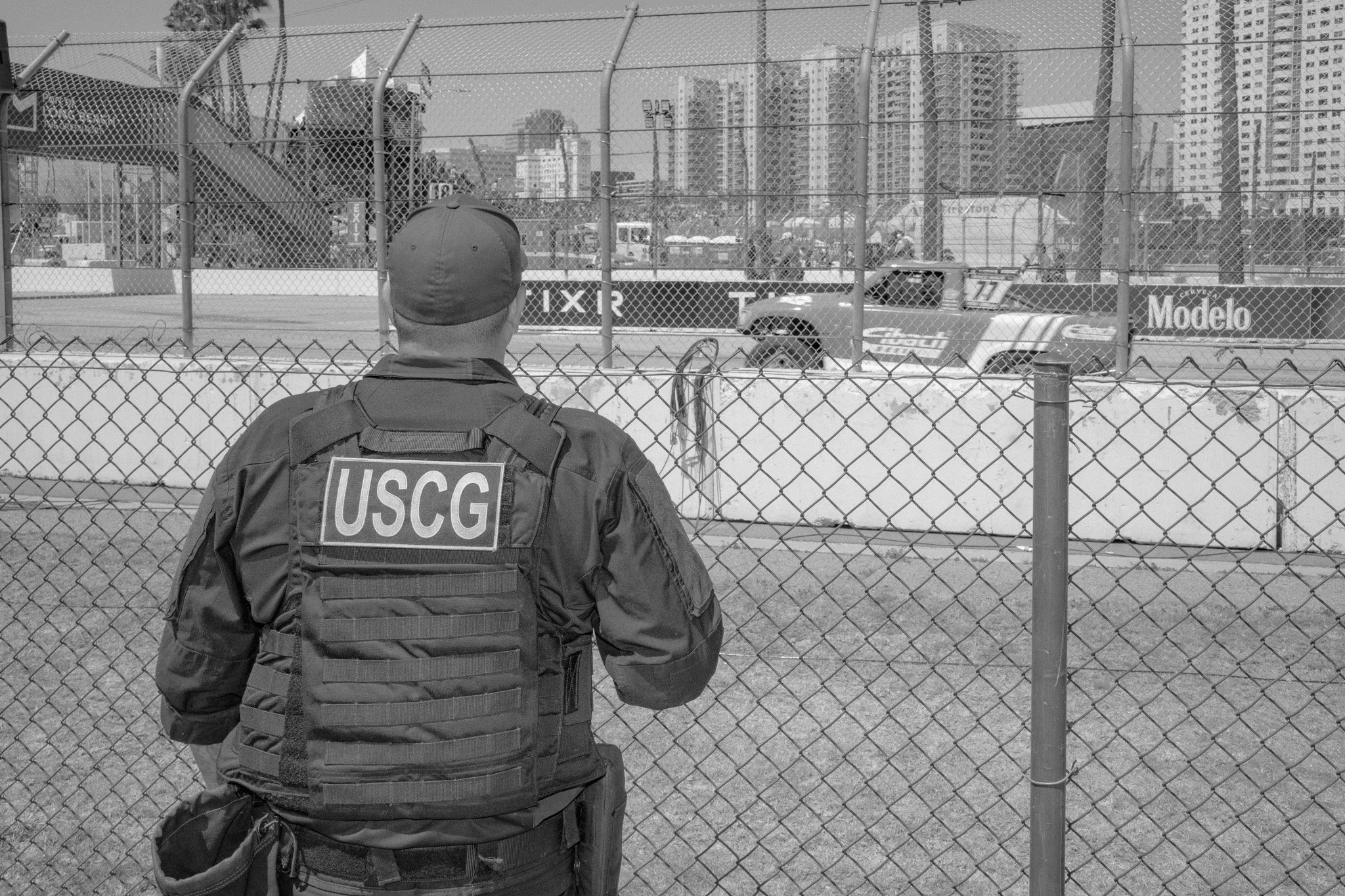 USCG officer standing in front of a chain-link fence watching a race track with a pickup truck passing by.