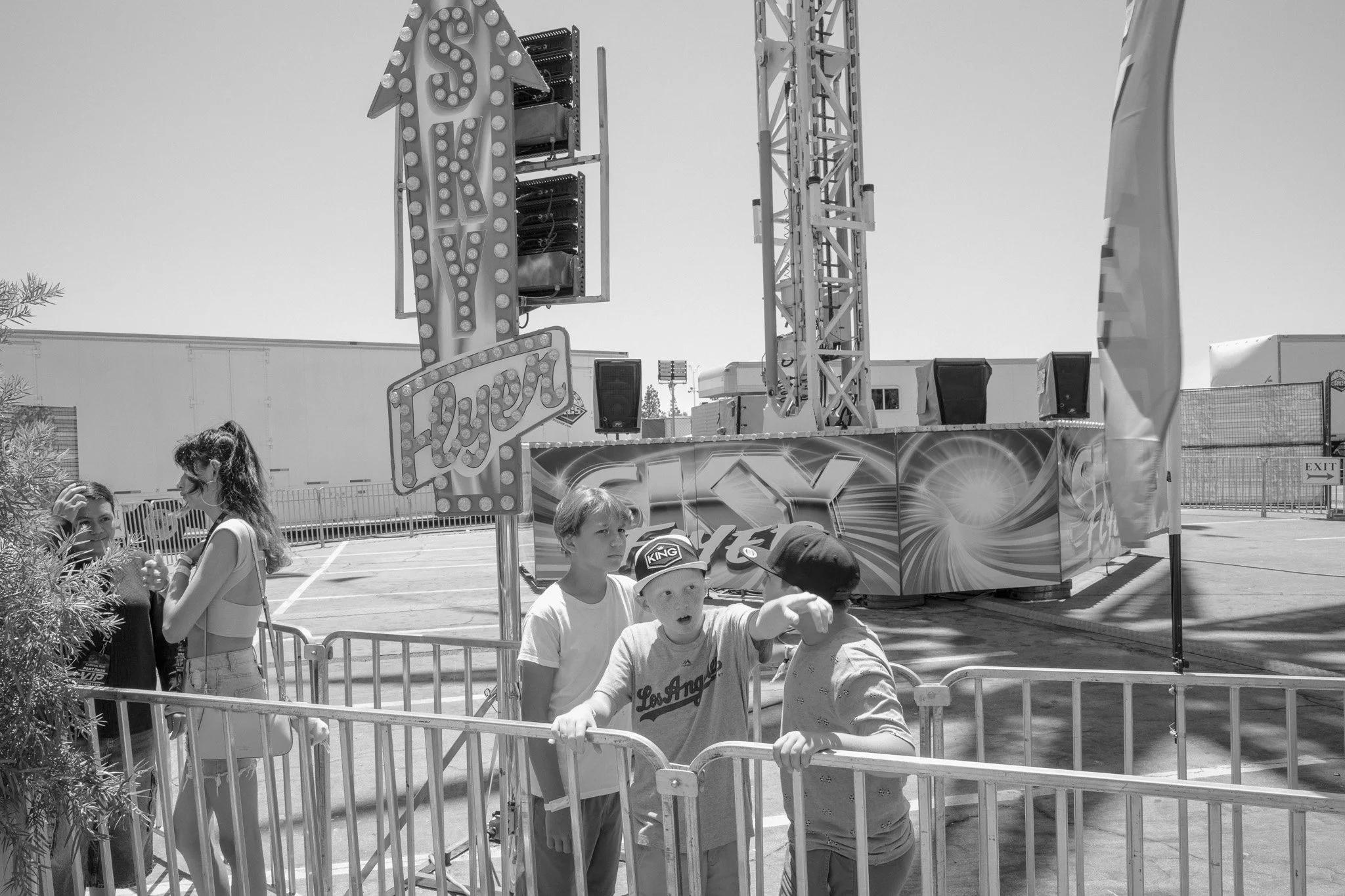 A group of children standing in front of a carnival ride with a large sign that says "SKY." The ride is tall with lights and a colorful backdrop, with some shadows cast on the ground.
