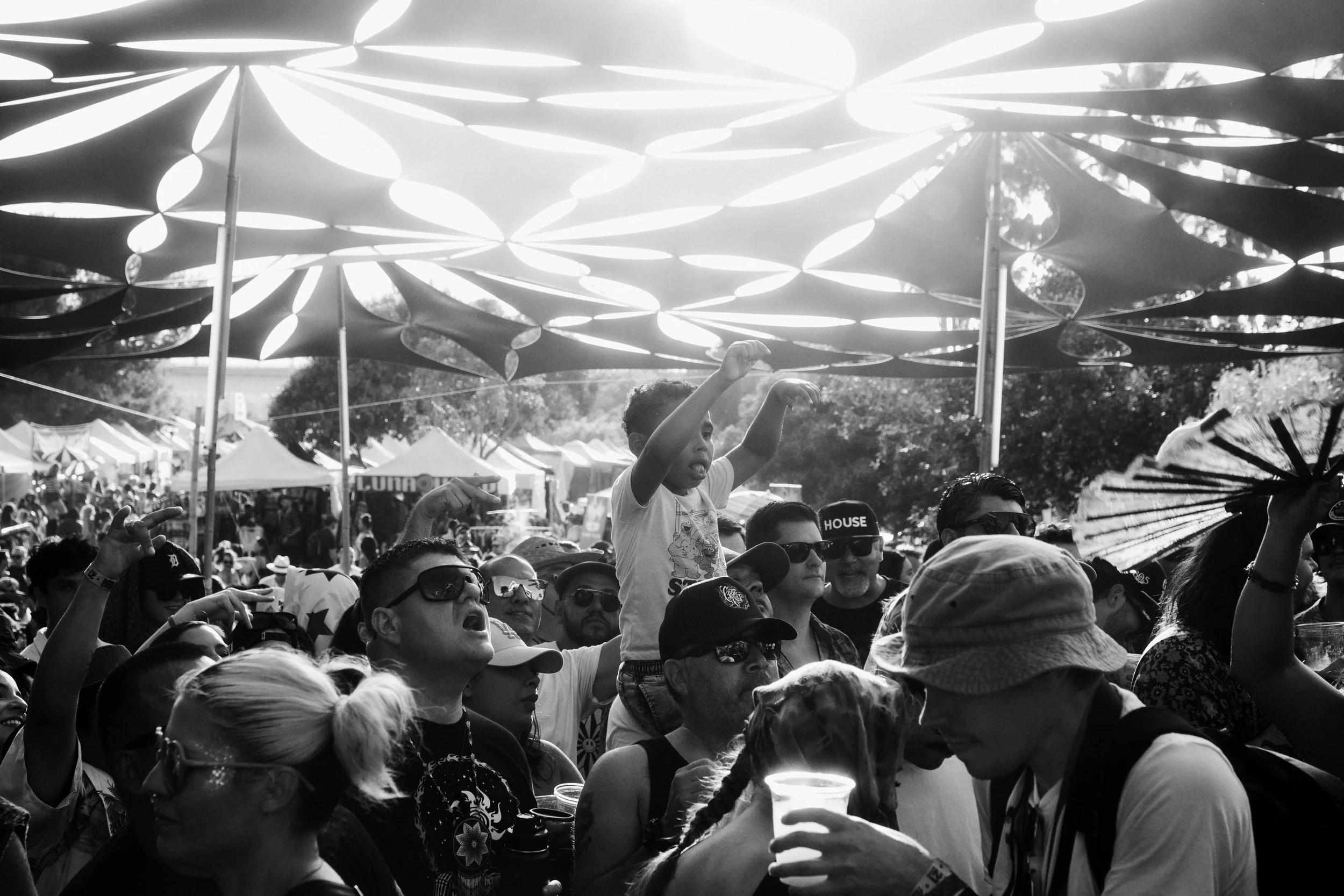 A black-and-white photo of a crowded outdoor event with many people, including children, under a decorative canopy. A boy is sitting on someone's shoulders, raising his arms, while others are wearing sunglasses and hats. Tents are visible in the back